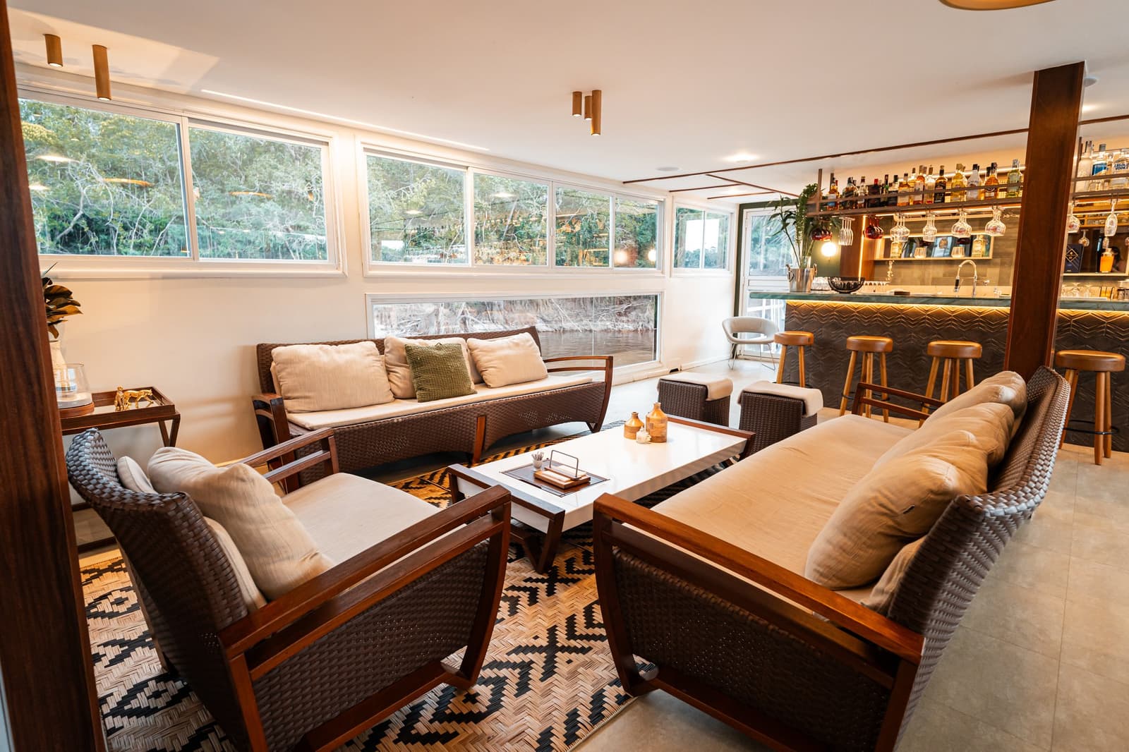 Ein Sundowner, die Bar griffbereit, der Regenwald direkt vor dem Fenster. Gemütliche Lounge mit Korbmöbeln, cremefarbenen Kissen und gemustertem Teppich auf einer Luxus-Amazonas-Kreuzfahrt in Brasilien, mit vollständig bestückter Bar und Panoramafenstern zum Fluss und Regenwald