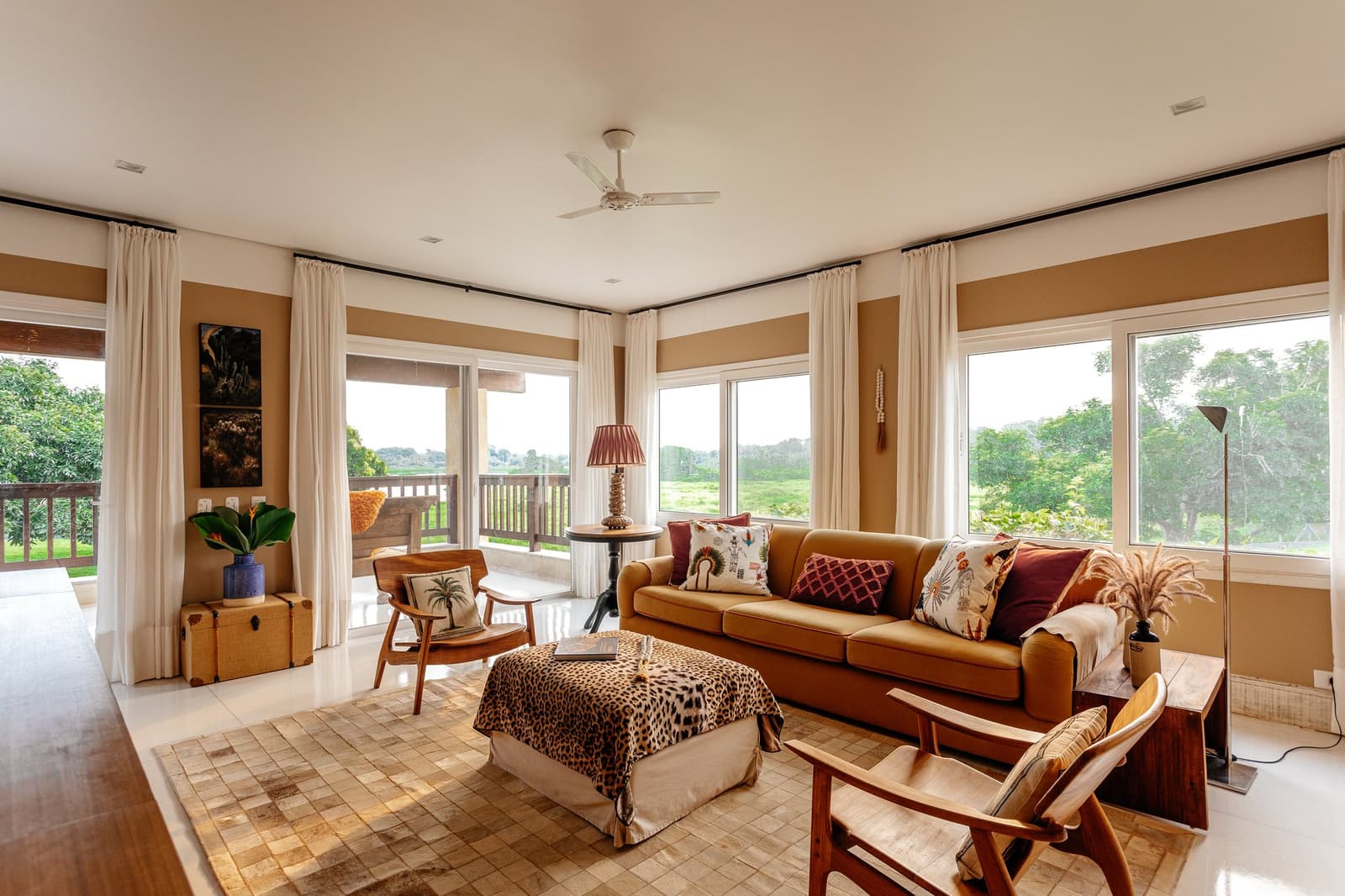Warm earth tones, wild views, and a ceiling fan doing honest work. Photo: Felipe Castellari Bright lodge living room on a luxury Pantanal and Amazon trip, featuring a caramel sofa with printed cushions, a leopard-print ottoman, timber armchairs, and floor-to-ceiling windows overlooking open wetland and green treeline