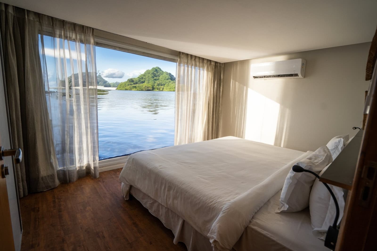 Einschlafen mit dem Fluss, aufwachen mit dem Wald. So sieht ein guter Morgen aus. Klimatisierte Kabine auf einem Luxus-Amazonaskreuzfahrtschiff in Brasilien mit Doppelbett, Holzboden und raumhohen Fenstern mit Blick auf ruhiges dunkles Flusswasser und eine bewaldete Insel