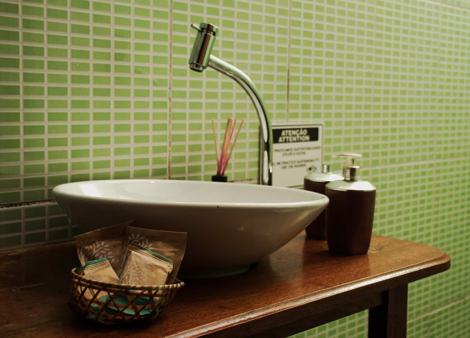 Even the soap dispenser is making a point about sustainability. White vessel sink on a dark wood vanity with green mosaic tiles, a small wicker basket of toiletry sachets, and a bilingual sustainability notice on the wall — part of an amazon and pantanal tour lodge bathroom