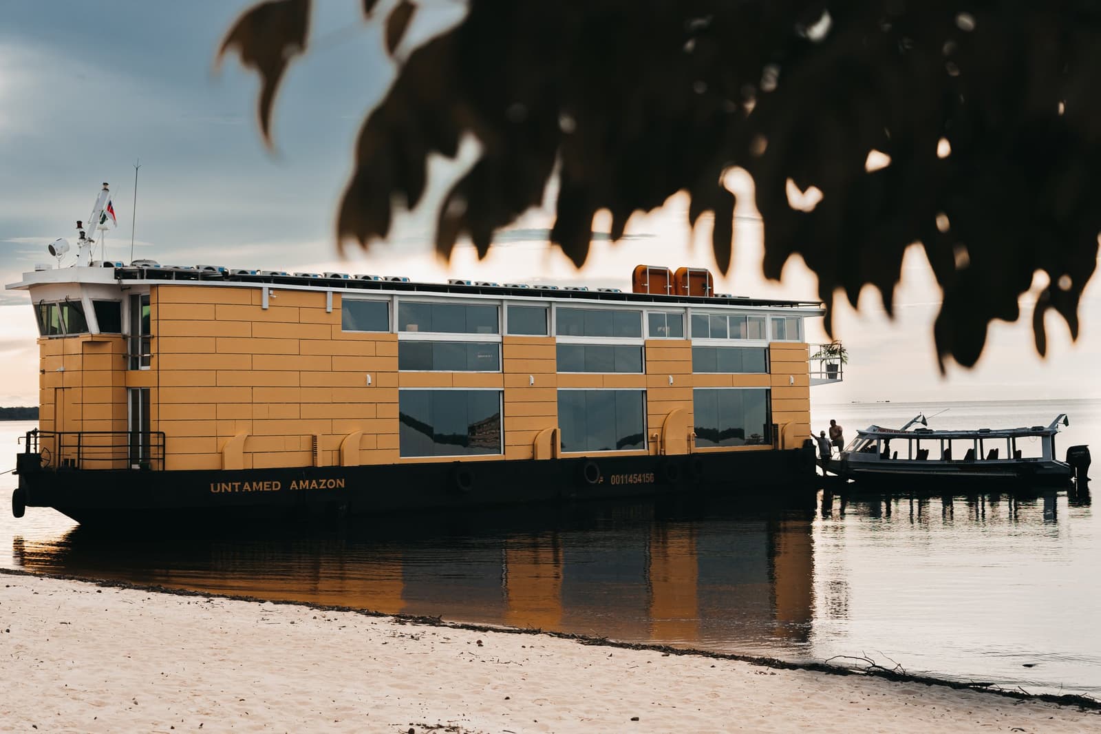 Fünf Tage, ein Fluss, eine Basis, der Rest ergibt sich von selbst. Das Flusskreuzfahrtschiff Untamed Amazon, ein gelbes, solarbetriebenes Schiff für Luxuskreuzfahrten im brasilianischen Amazonas, liegt in der Abenddämmerung an einem Sandstrand, flankiert von einem kleinen Beiboot und Ästen im Vordergrund