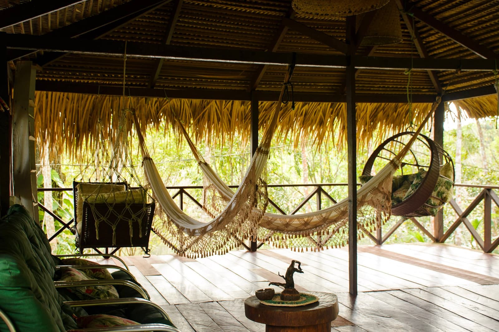 Pick a hammock, any hammock. Harder than it sounds. Open-sided jungle lodge terrace with multiple fringed hammocks, a wicker hanging chair, and a thatched palm roof overlooking dense forest greenery — the kind of rest stop an amazon and pantanal tour earns you