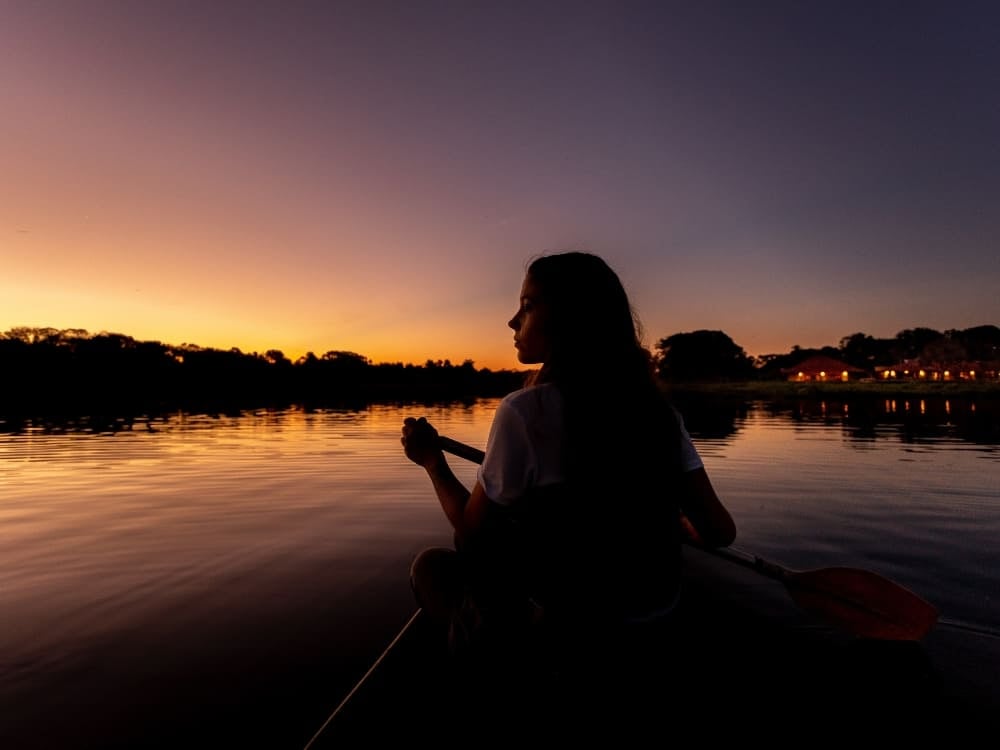 Eine Frau paddelt bei Sonnenuntergang in einem Kanu auf einem ruhigen Fluss, was die friedliche Natur unterstreicht (Pantanal, Brasilien). Eine Frau paddelt bei Sonnenuntergang in einem Kanu auf einem ruhigen Fluss, was die friedliche Natur unterstreicht (Pantanal, Brasilien).