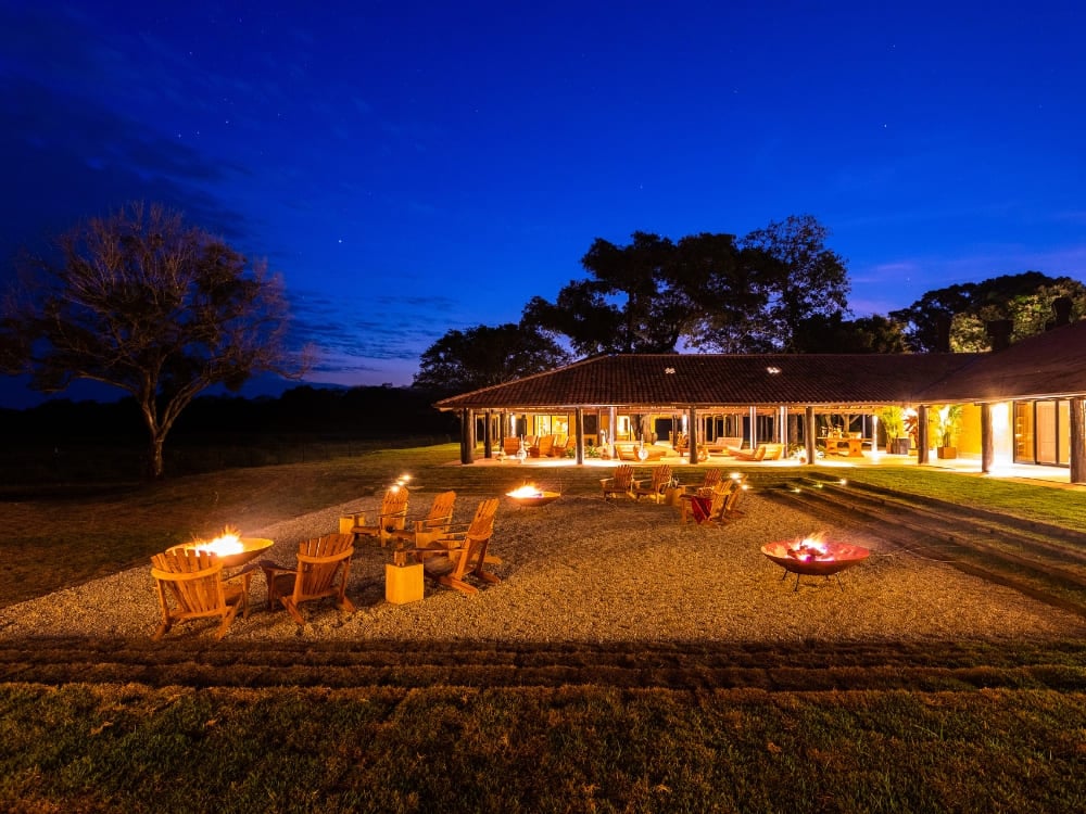 Der Außenbereich der Casa Caiman bei Nacht, mit Feuerstellen und Holzstühlen unter einem tiefblauen Sternenhimmel im Pantanal. Der Außenbereich der Casa Caiman bei Nacht, mit Feuerstellen und Holzstühlen unter einem tiefblauen Sternenhimmel im Pantanal.