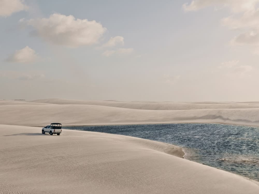 . A white 4x4 tour vehicle driving across vast white sand dunes next to a bright blue lagoon.
