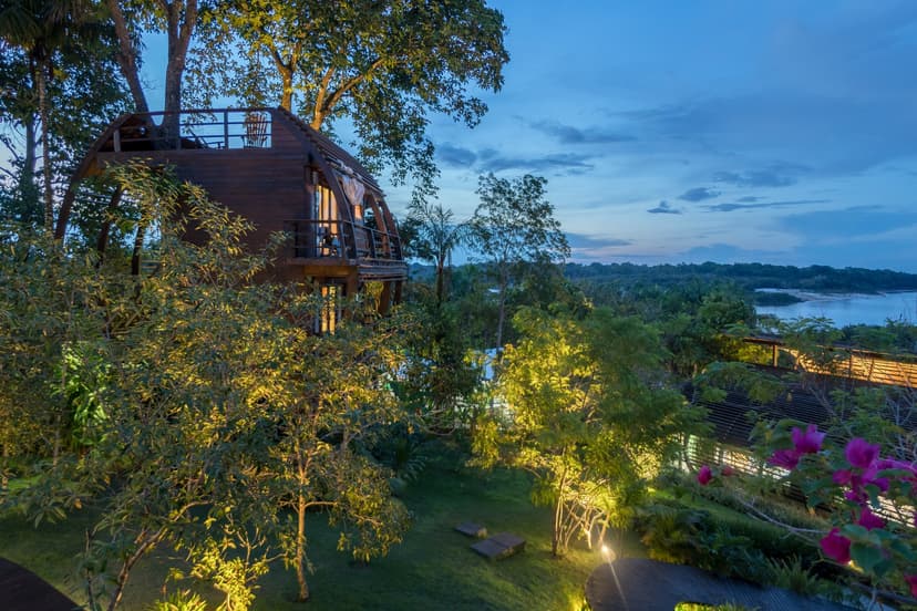 A curved timber treehouse bungalow at Mirante do Gavião glows with warm interior light at dusk, surrounded by lush tropical garden and overlooking a wide river bend under a blue twilight sky A curved timber treehouse bungalow at Mirante do Gavião glows with warm interior light at dusk, surrounded by lush tropical garden and overlooking a wide river bend under a blue twilight sky