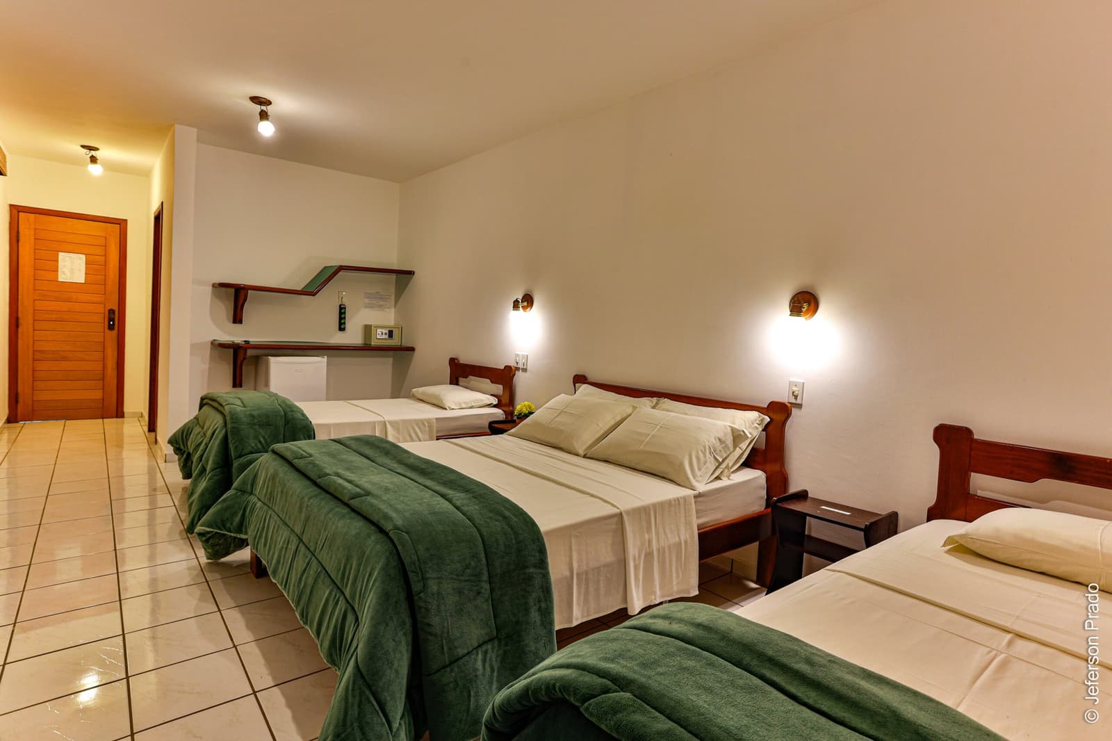Clean, comfortable, and ready for an early wake-up call. Triple guest room on the Pantanal Jaguar Photography Tour, with three wooden-framed beds dressed in white linen and green blankets, warm wall sconces, tiled floor, and a small desk area near the entrance corridor