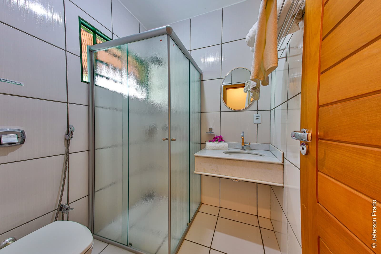 Small, tidy, and ready for you after a long day in the field. Clean private bathroom with glass-enclosed shower cabin, white tiled walls, vanity with round mirror, fresh towels and a small floral decoration, at accommodation on the Pantanal Jaguar Photography Tour