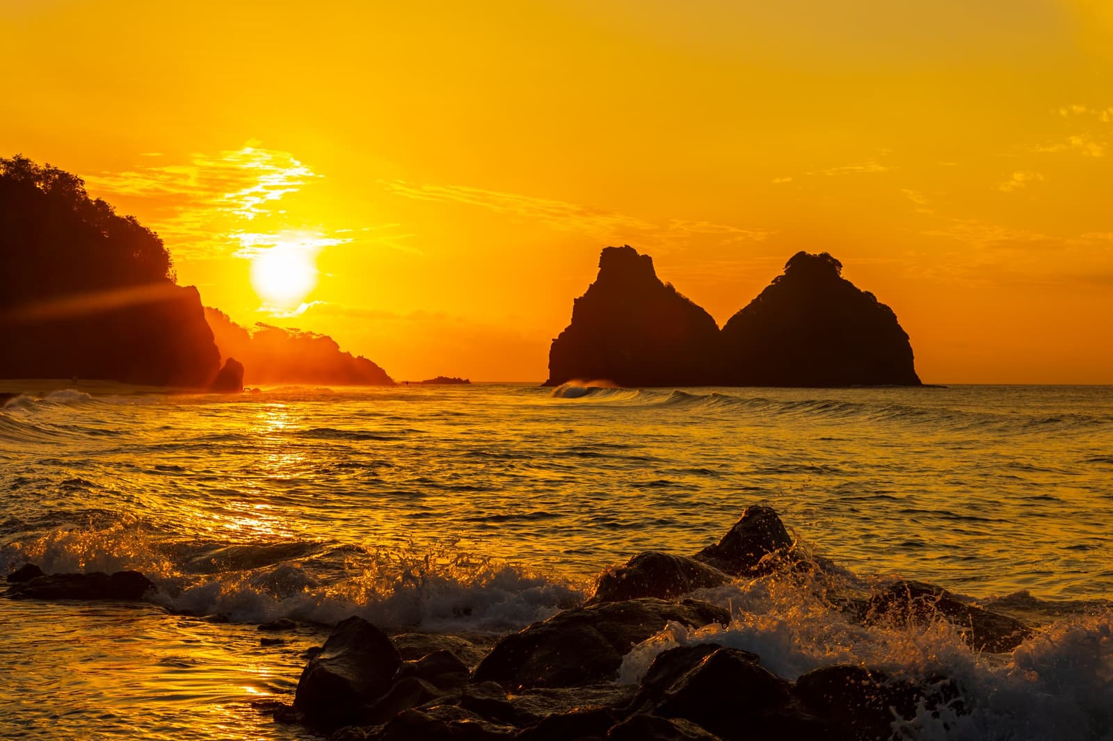 Molten gold poured across the Atlantic — the island's dramatic silhouette at day's end. Photo: Reuber Duarte Fiery golden sunset over silhouetted rocky sea islets off Fernando de Noronha, waves crashing over foreground boulders, part of a 2-Day Diving Package in Fernando de Noronha