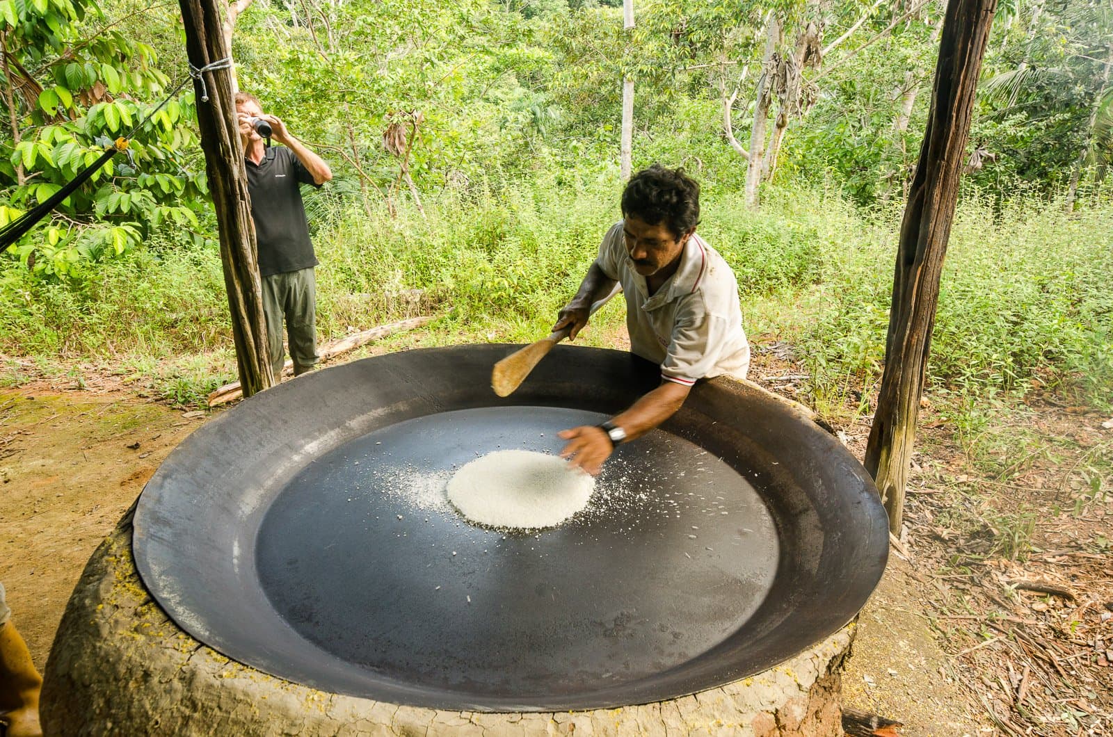 Cassava flour, hand-stirred on a scorching iron plate — a staple food tradition alive in the riverside community. Photo: Leonide Principe A local man toasts cassava flour on a large iron griddle over an open fire during an amazon rainforest ecolodge trip in brazil, with a visitor photographing birds in the jungle background