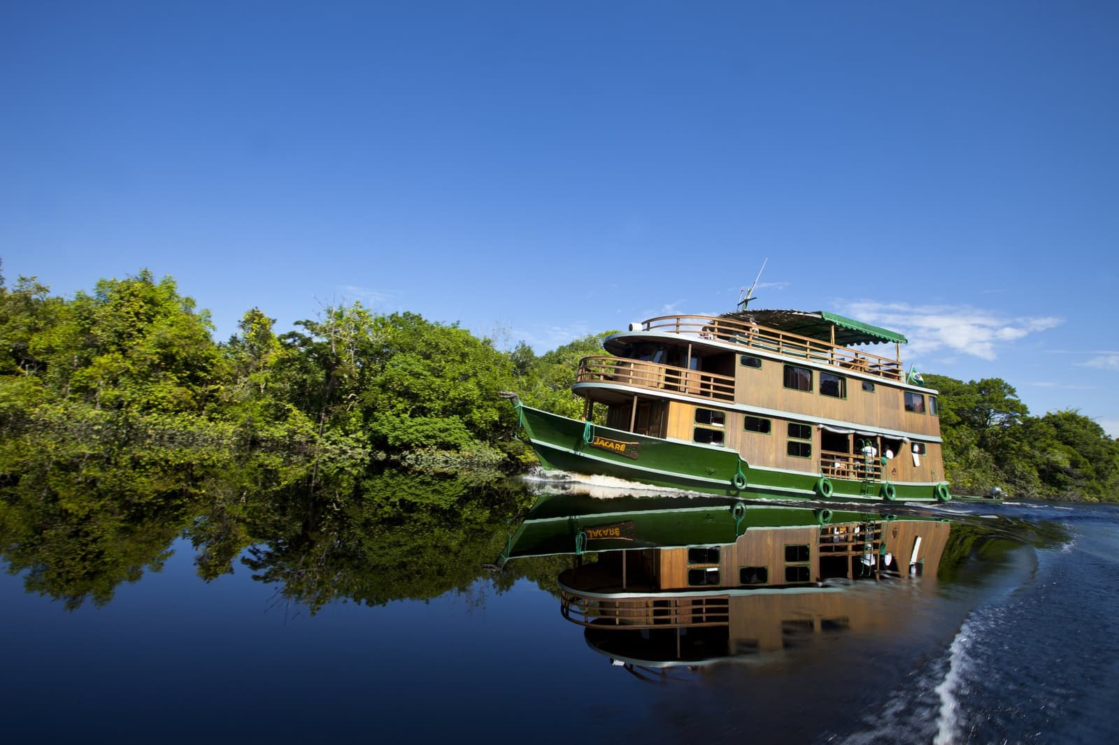 Three decks, open railings, and a river that doubles the sky — the Jacaré sets off into the heart of the Amazon. The Jacaré houseboat cruising a glassy dark-water river on an Amazon River Cruise Brazil, its green-and-timber hull perfectly mirrored in the still water with dense rainforest lining the banks