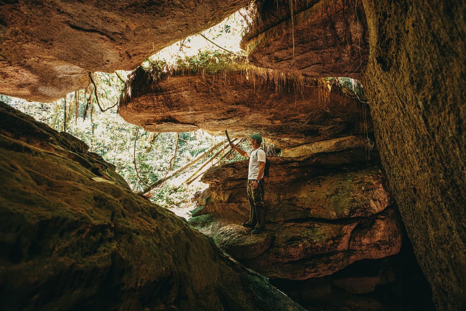 Hier heeft de natuur miljoenen jaar aan gebouwd, zonder blauwdruk. Een gids staat in de oeroude zandsteenformaties van de Madadá-grotten tijdens een trektocht vanuit Anavilhanas Jungle Lodge Brazil, terwijl hij omhoogkijkt naar het dramatische overhangende rotsplafond met hangende wortels