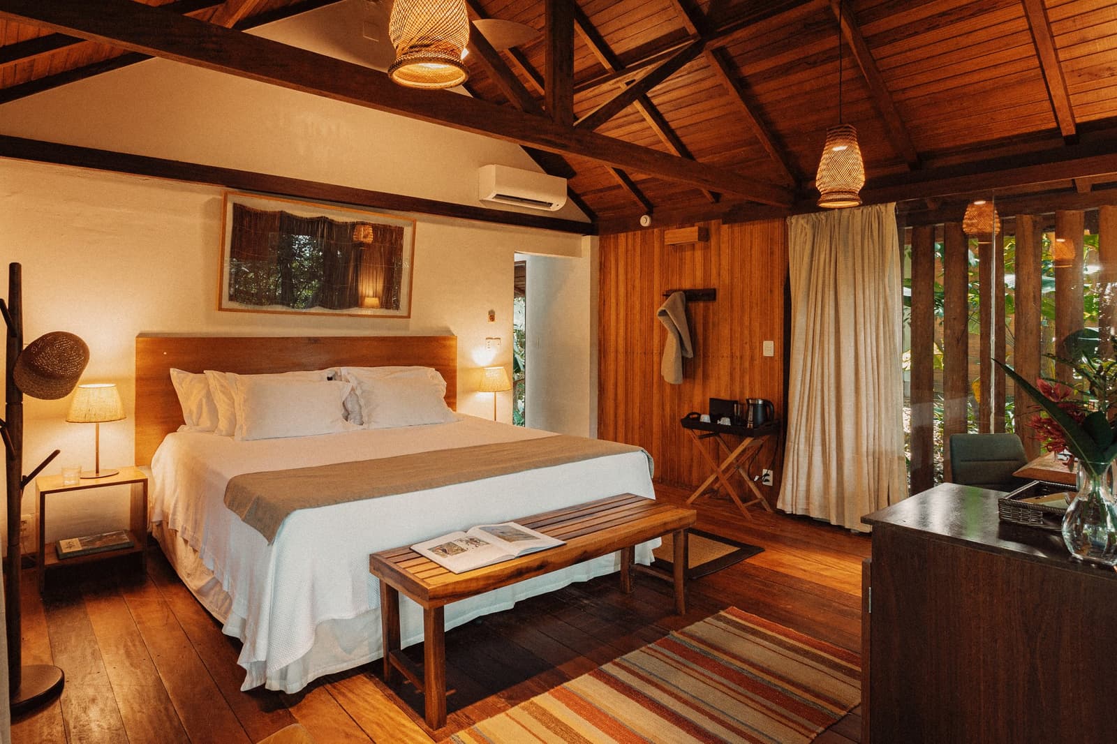 Na een dag op de rivier is dit precies de plek om op adem te komen. Sfeervol verlichte chaletskamer in Anavilhanas Jungle Lodge Brazil met een kingsize bed met witte beddengoed, donkere houten dakbalken, gevlochten rotan hanglampen en een raam met uitzicht op weelderig groen