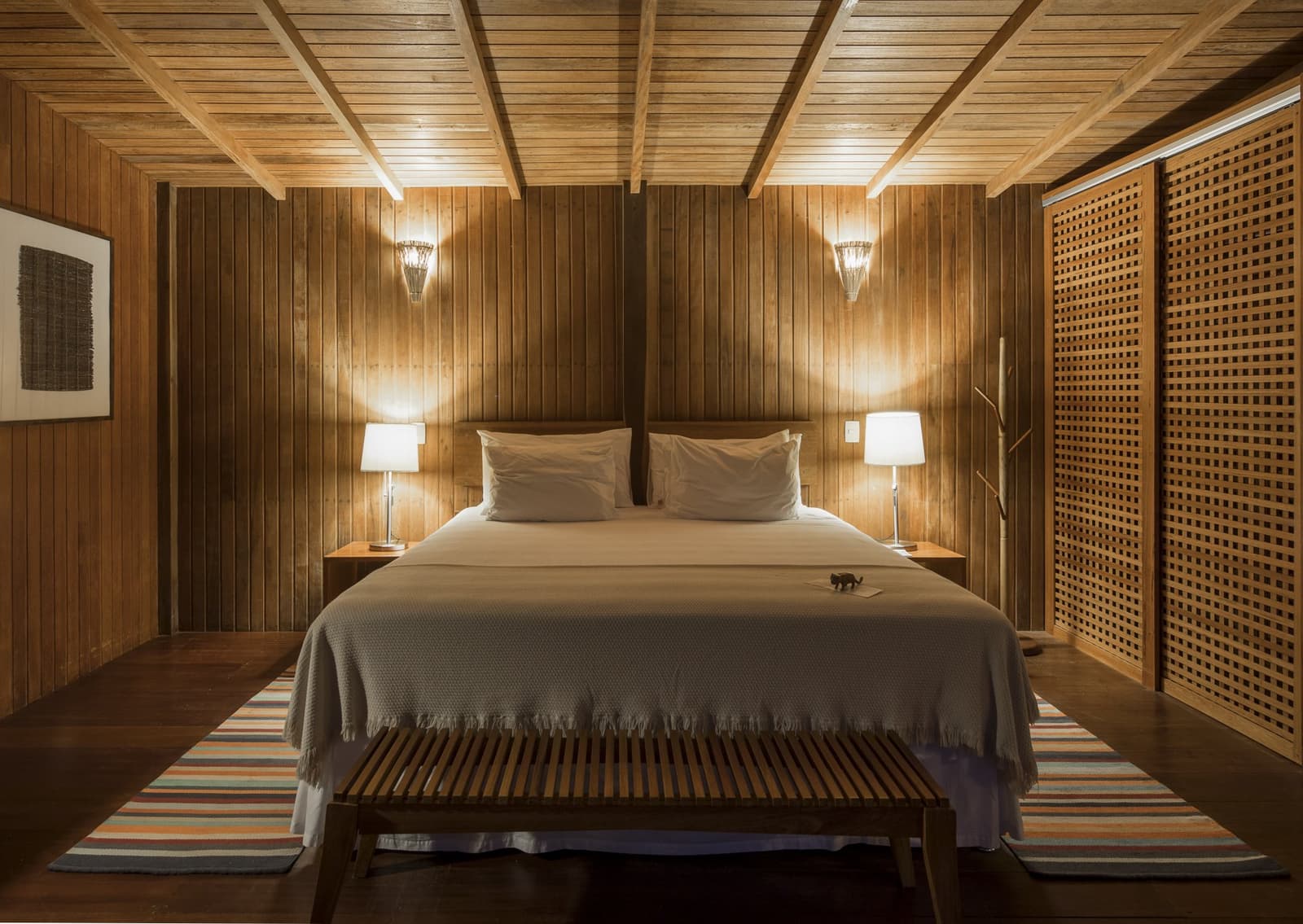 Hout van vloer tot plafond, het oerwoud op een steenworp afstand. Warm verlichte tweepersoonskamer van Anavilhanas Jungle Lodge Brazil met houten lambrisering op wanden en plafond, een kingsize bed met witte beddengoed en grijze franjedeken, twee nachtkastlampen, houten traliewerk en een gestreept vloerkleed op donker parket