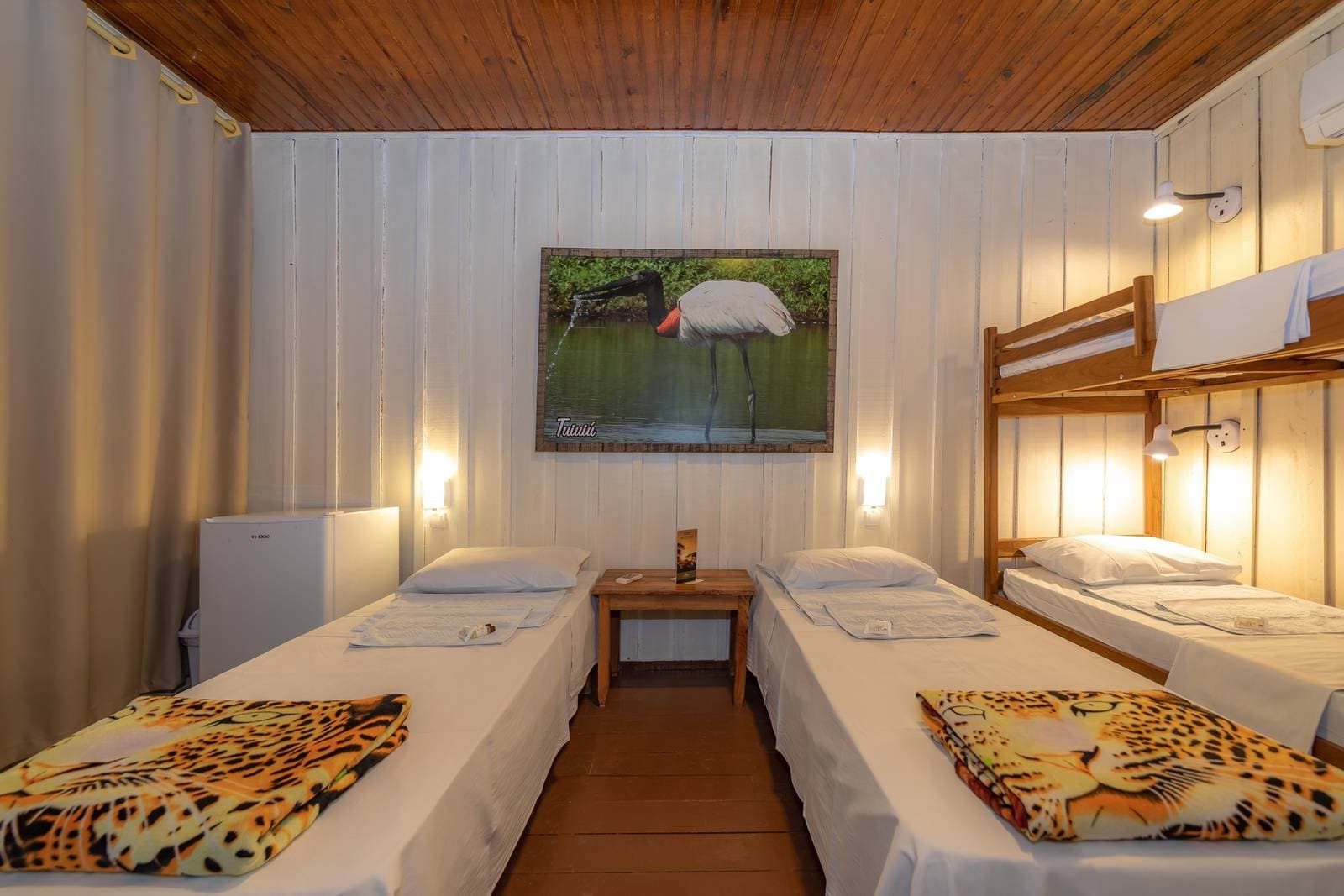 Jaguar blankets, warm lamplight, and a jabiru on the wall — simple comforts after a day in the wetlands. Cozy multi-bed lodge room on a budget pantanal tour, featuring two single beds with jaguar-print blankets, a wooden bunk bed, warm wall lighting, and a framed jabiru stork artwork above a small bedside table