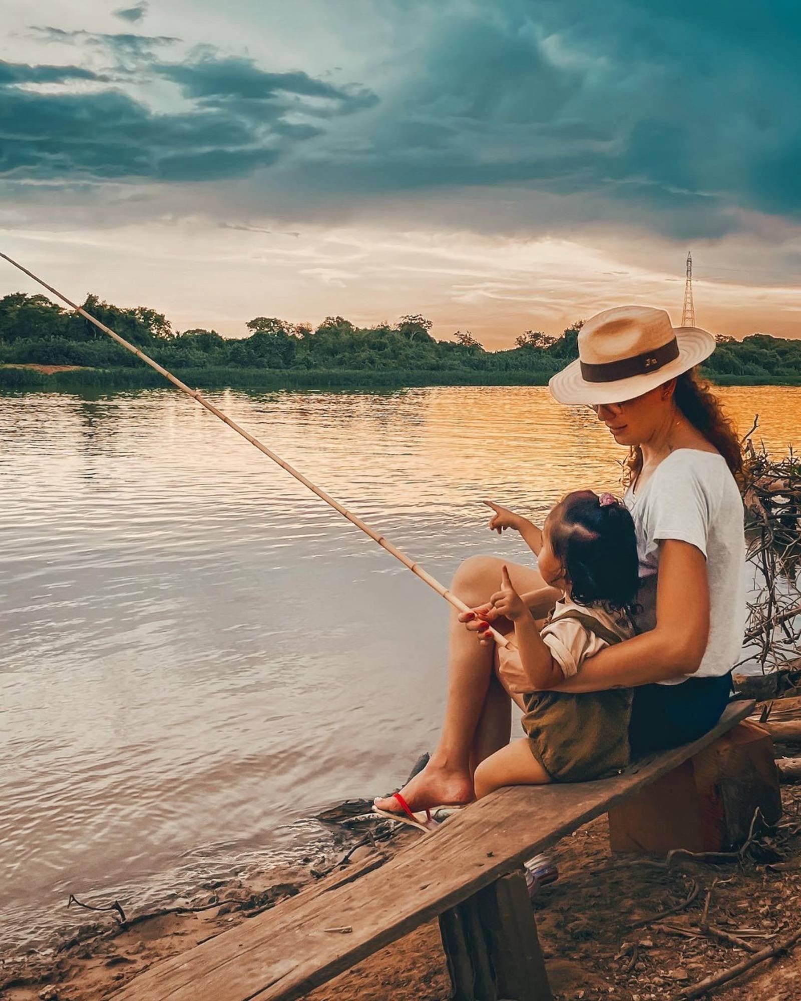 Bamboo rod, tiny hands, and a river turning gold — piranha fishing as it should be. A woman in a panama hat holds a young child on her lap while fishing with a bamboo rod on a calm river at sunset during a budget Pantanal tour, golden light reflecting off the water