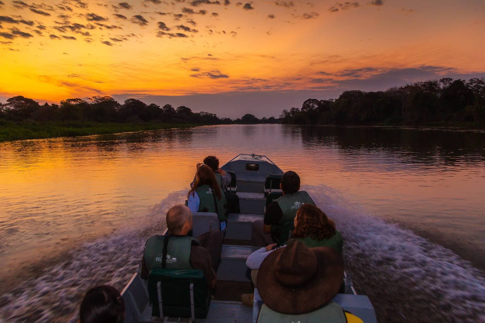 Golden hour on the river — every traveller's favourite moment. Travellers in life jackets aboard a motorboat on a calm river at sunset during a budget Pantanal tour, with a vivid orange sky reflecting on the water and silhouetted jungle on both banks