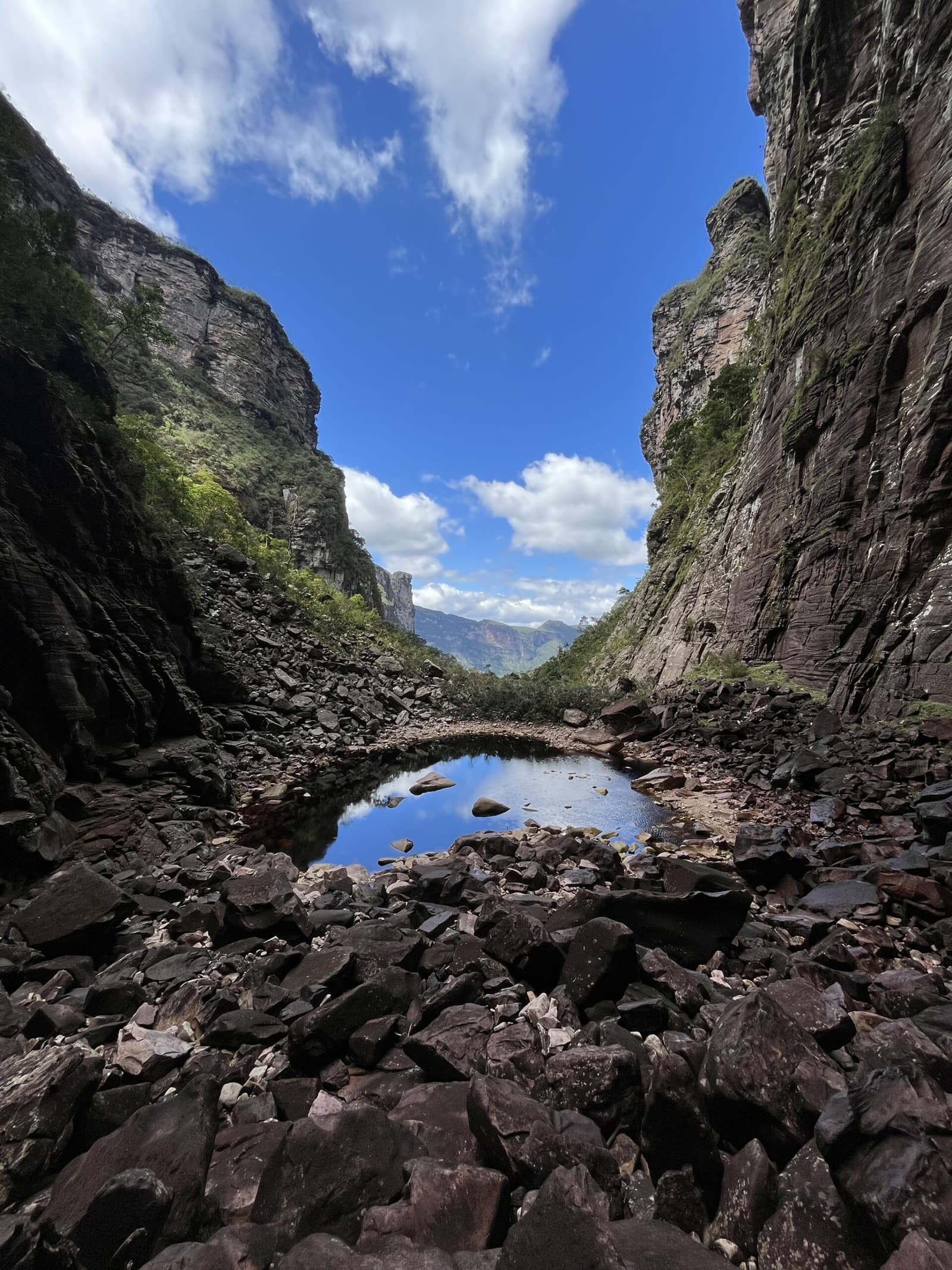 Sky caught in stone — a quiet pool between canyon walls. Photo: Lucas Neves A still rockpool mirrors the blue sky between towering sandstone canyon walls on the Chapada Diamantina trek, with scattered boulders leading toward open mountains in the distance