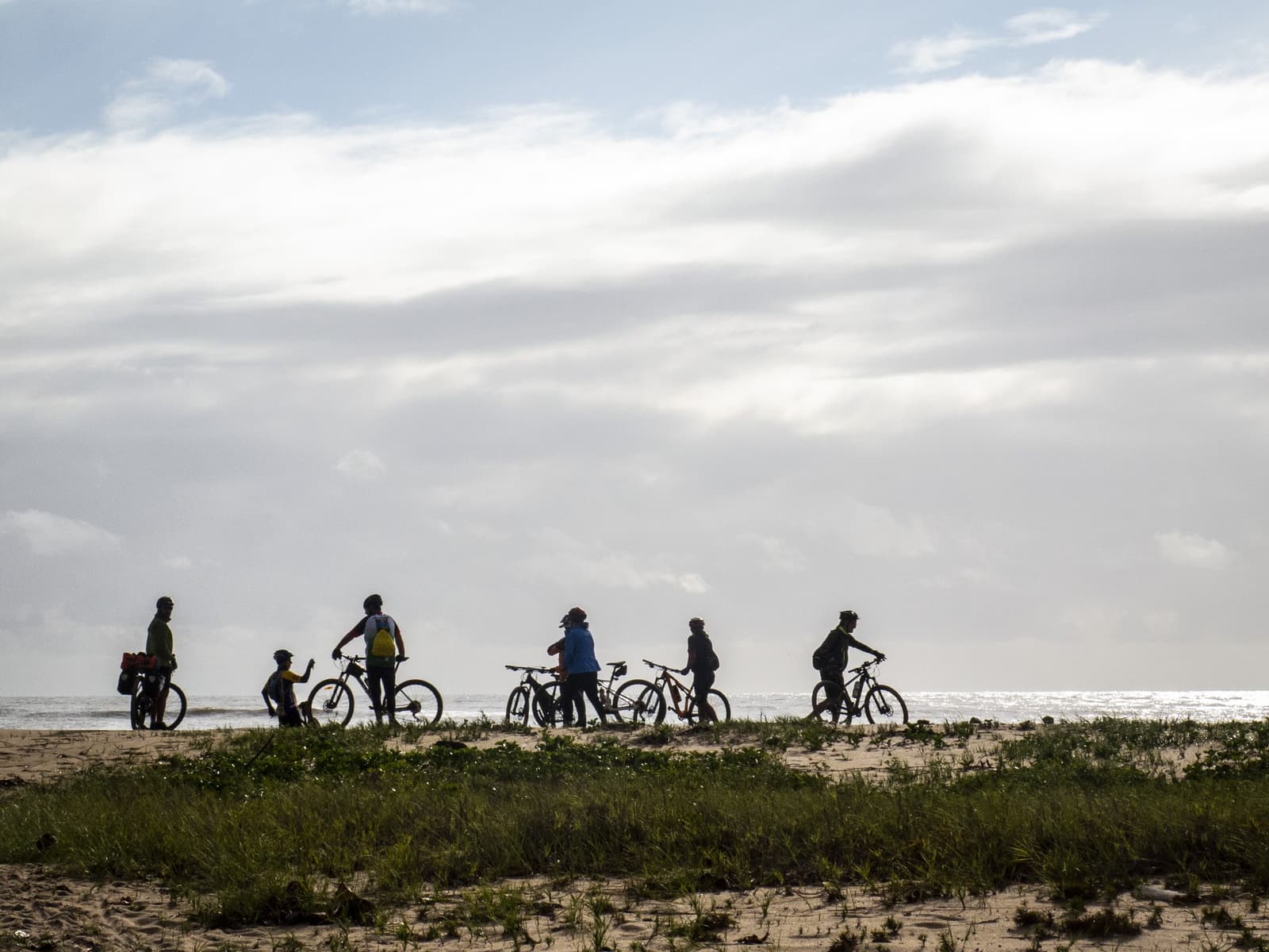 Even pauzeren op het zand — de kust van Bahia strekt zich eindeloos uit. Silhouetten van vijf fietsers met mountainbikes die pauzeren op een kustduin met lage begroeiing, met de glinsterende Atlantische Oceaan op de achtergrond — een moment tijdens het fietsen in Bahia tussen Ilhéus en Itacaré