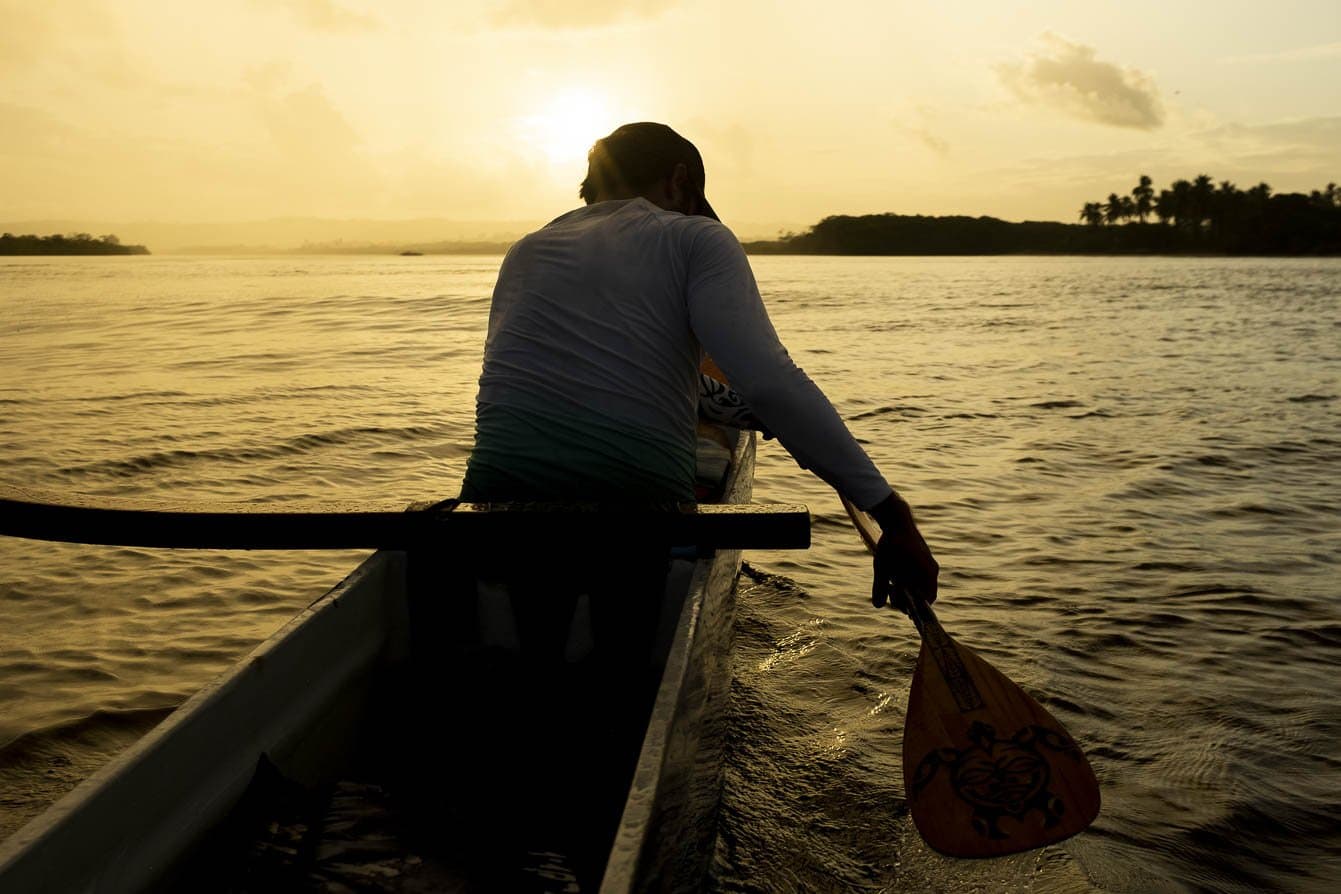 Gouden uur op de Rio das Contas — peddelen richting de Bahiaanse zonsondergang. Een peddellaar als silhouet tegen een gouden zonsondergang bestuurt een Hawaïaanse kano over de rustige Rio das Contas bij Itacaré, tijdens een fietsvakantie in Bahia