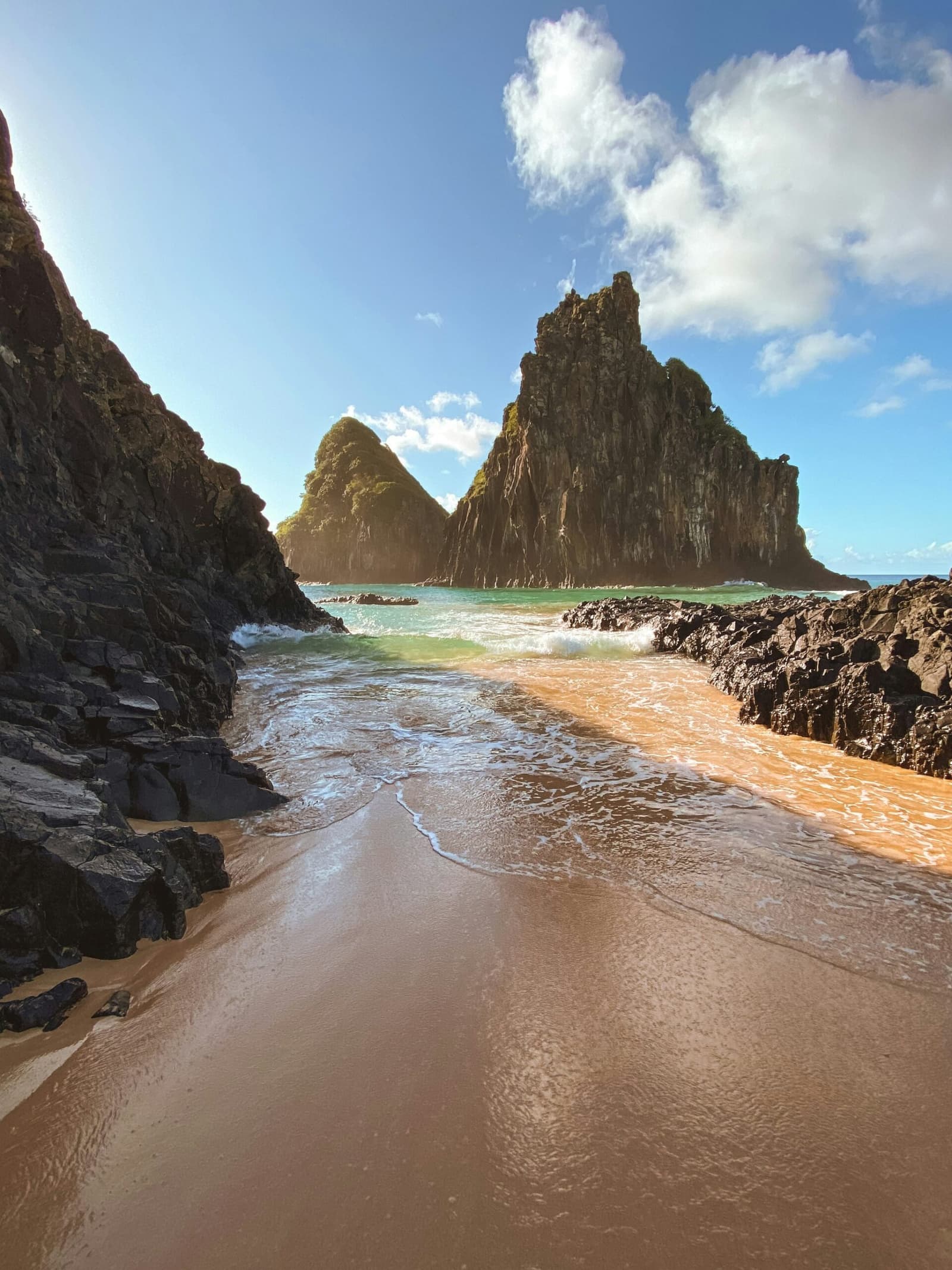 Dit soort hoekjes houdt Fernando de Noronha klein en bijzonder. Een smalle goudgekleurde zandbaai omgeven door donker vulkanisch gesteente, met twee imposante bemoste rotspieken die oprijzen boven de turquoise Atlantische oceaan