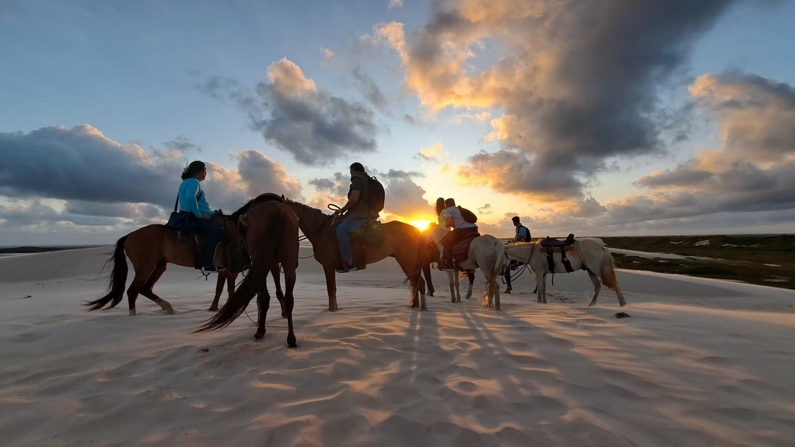 Riders paused at golden hour, shadows stretching long across the dunes. A group of riders on horseback silhouetted against a blazing sunset over the white sand dunes of Lençóis Maranhenses, with dramatic clouds spreading across the sky during a dune crossing on horseback