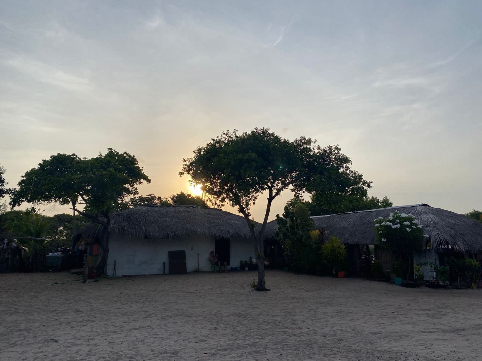 O fim de tarde que chega devagar e convida a ficar. Casas caiadas com telhados de palha e uma árvore solitária em silhueta contra o pôr do sol sobre chão de areia, uma comunidade local típica ao longo do passeio a cavalo nos Lençóis Maranhenses
