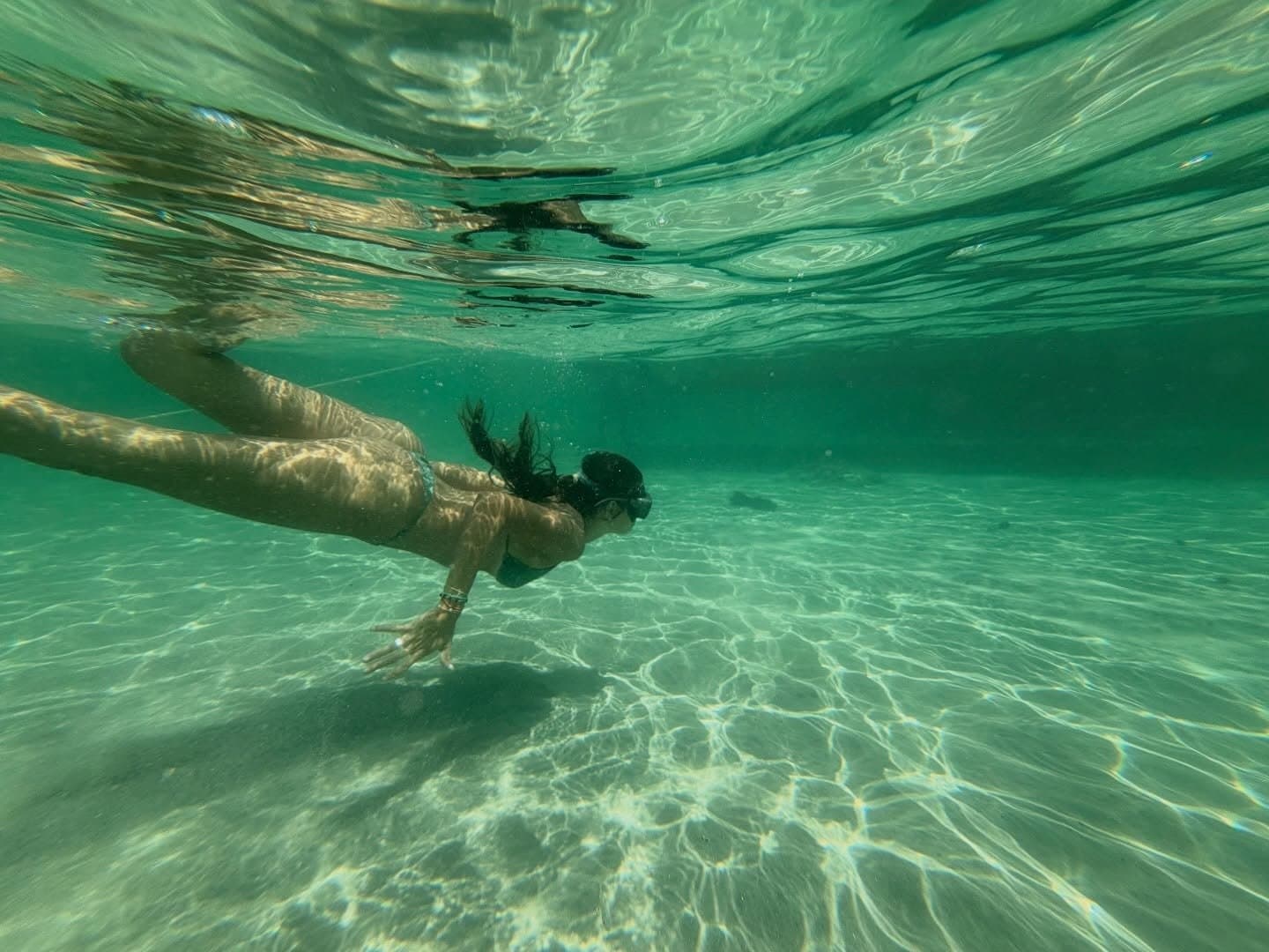 Pure emerald water, sunlight rippling down — every lagoon is an invitation. A woman free-diving through crystal-clear turquoise water above a sandy bottom, captured underwater during a Lençóis Maranhenses hike swim stop