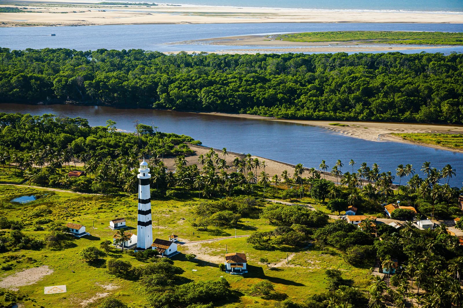 A striped sentinel above the river, palms, and dunes beyond. Photo: Gabriel Boieras Aerial view of a tall black-and-white striped lighthouse surrounded by palm trees and green vegetation, with a wide river estuary and white sand dunes stretching to the horizon — a landmark on the Lençóis Maranhenses hike route