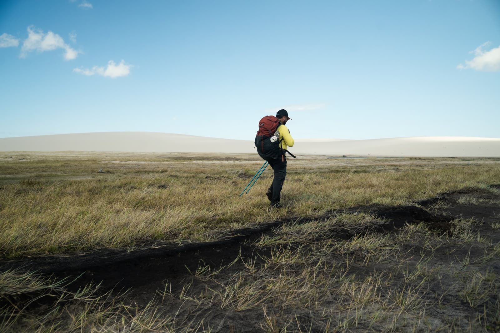 Poles out, pack loaded — the dunes are still a long way off. A trekker with a large red backpack and trekking poles crosses a wide flat grassland on the Lençóis Maranhenses hike, with pale sand dunes stretching across the horizon under a blue sky