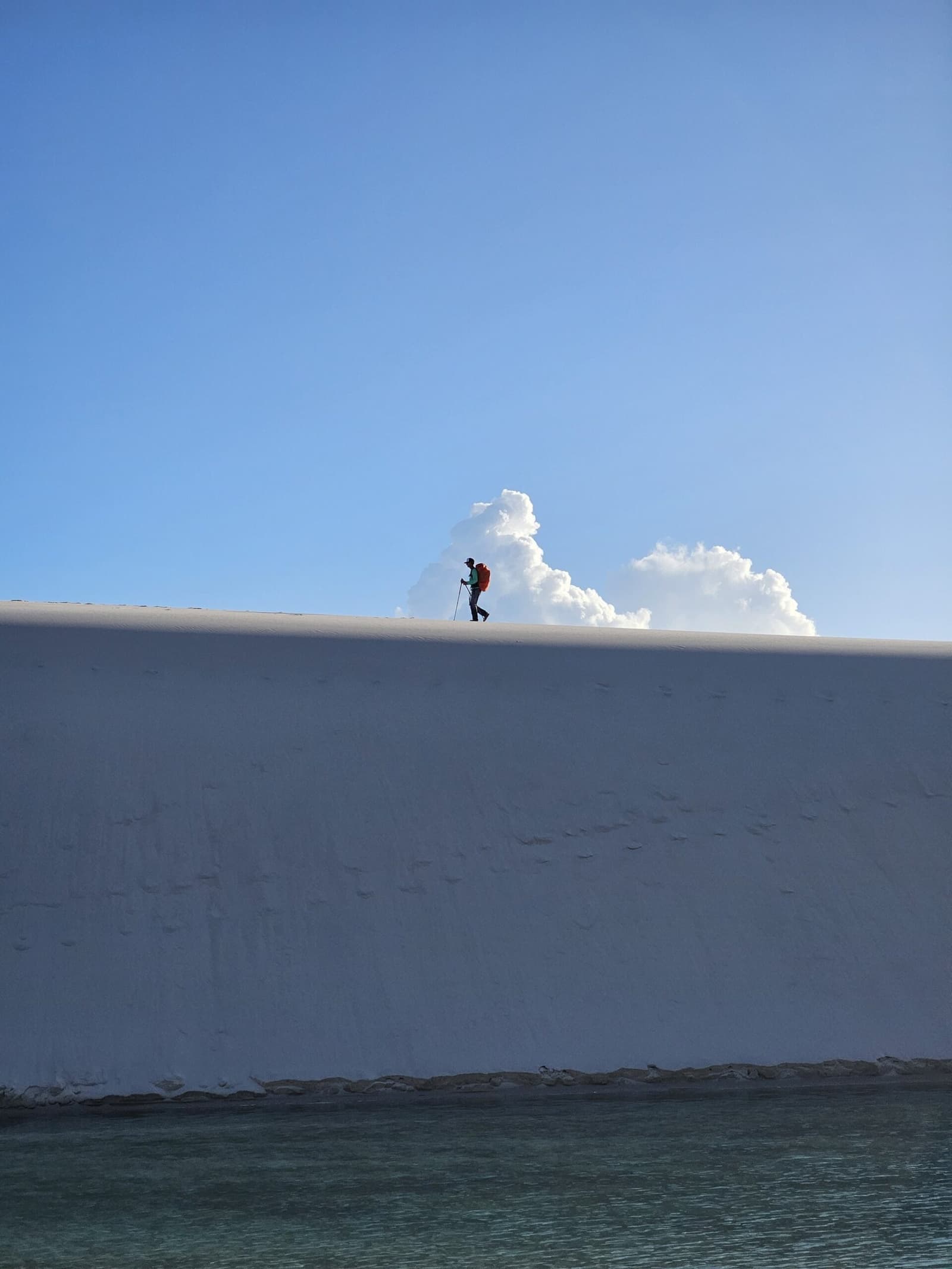 Small against the white — a single ridge between two worlds. A lone hiker with a red pack and trekking pole silhouetted on the razor-sharp crest of a brilliant white sand dune during the Lençóis Maranhenses hike, with turquoise lagoon water visible at the base and a towering cumulus cloud rising behind