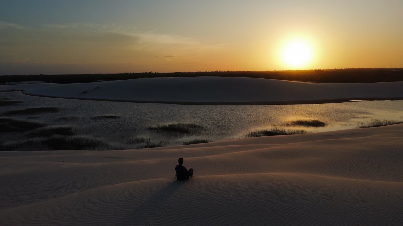 Quiet enough to hear the wind shift — sunset over the lagoons. A lone traveller sits silhouetted on white sand dunes overlooking a still lagoon at sunset in Lençóis Maranhenses Santo Amaro, with golden light reflecting off the water