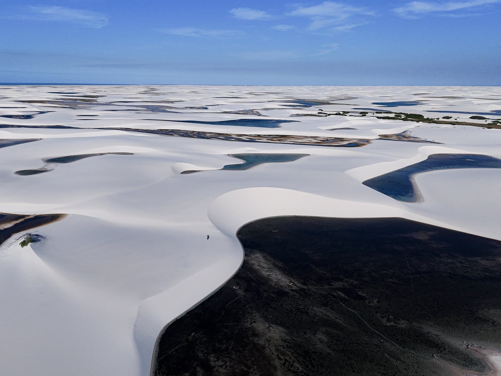Sculpted by wind and rain: an ocean of white dunes and hidden lagoons, reaching as far as the eye can see. Aerial view of the Lençóis Maranhenses Trek from Atins, showing vast white sand dunes stretching to the horizon with dark and blue freshwater lagoons nestled between them under a clear blue sky