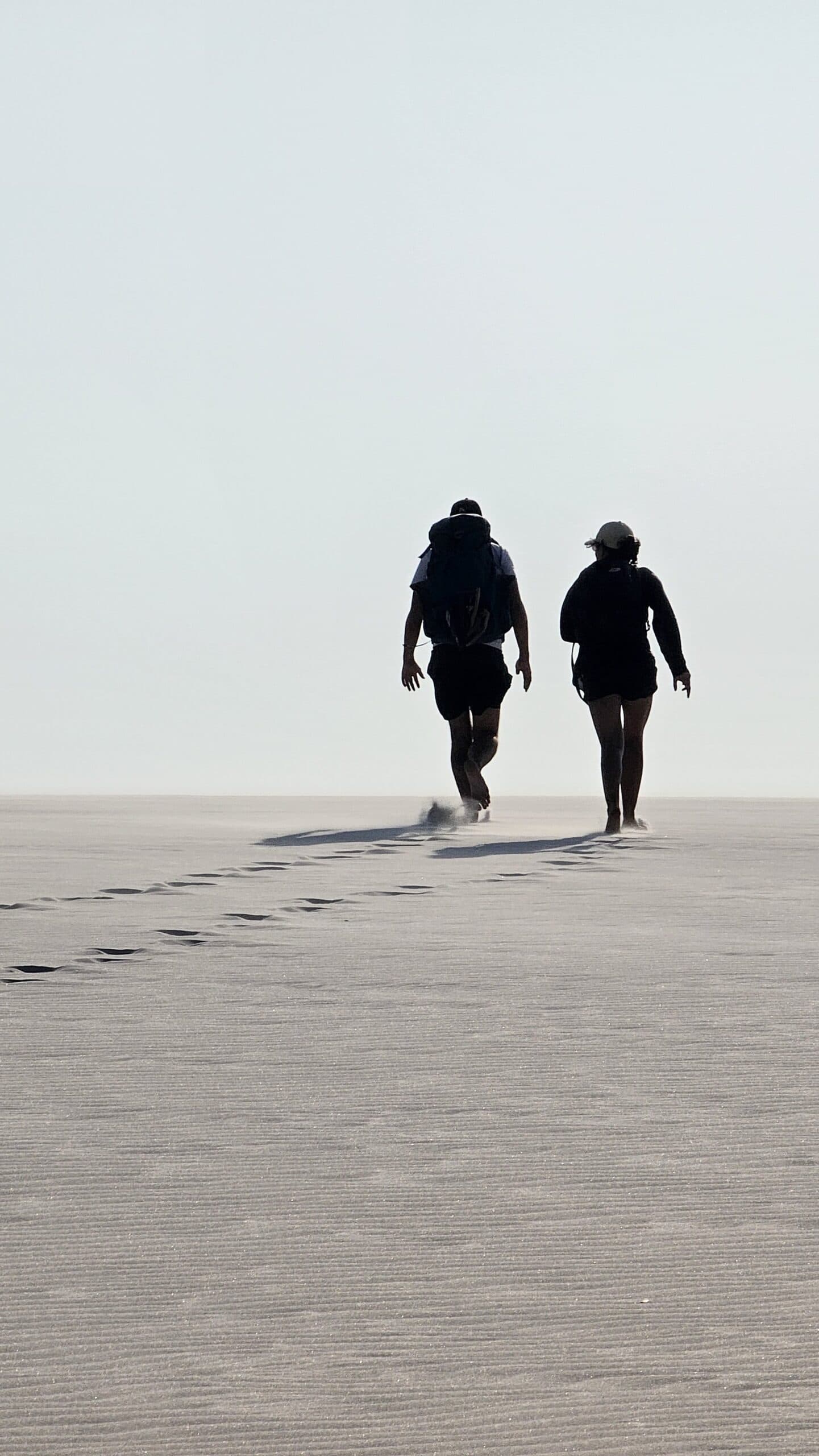 Footprints fading into the white — just you, your pack, and the dunes ahead. Two trekkers with backpacks walk barefoot across vast white sand dunes on the Lençóis Maranhenses trek from Atins, leaving a trail of footprints behind them under a pale hazy sky