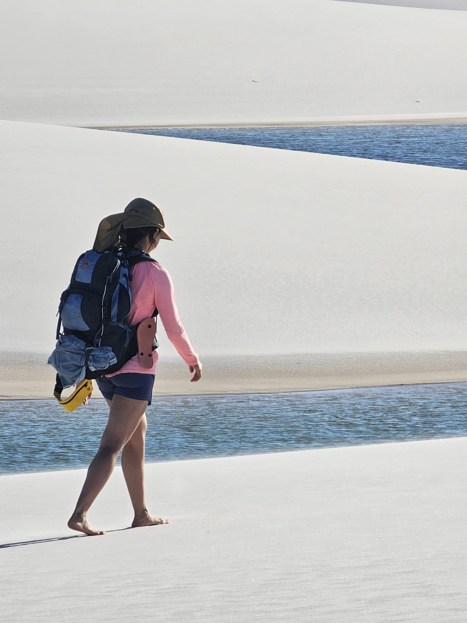 Barefoot, backpacked, and moving through one of the world's most surreal landscapes. A barefoot hiker with a backpack and sun hat walks along the edge of a freshwater lagoon between sweeping white sand dunes during a lencois maranhenses trek