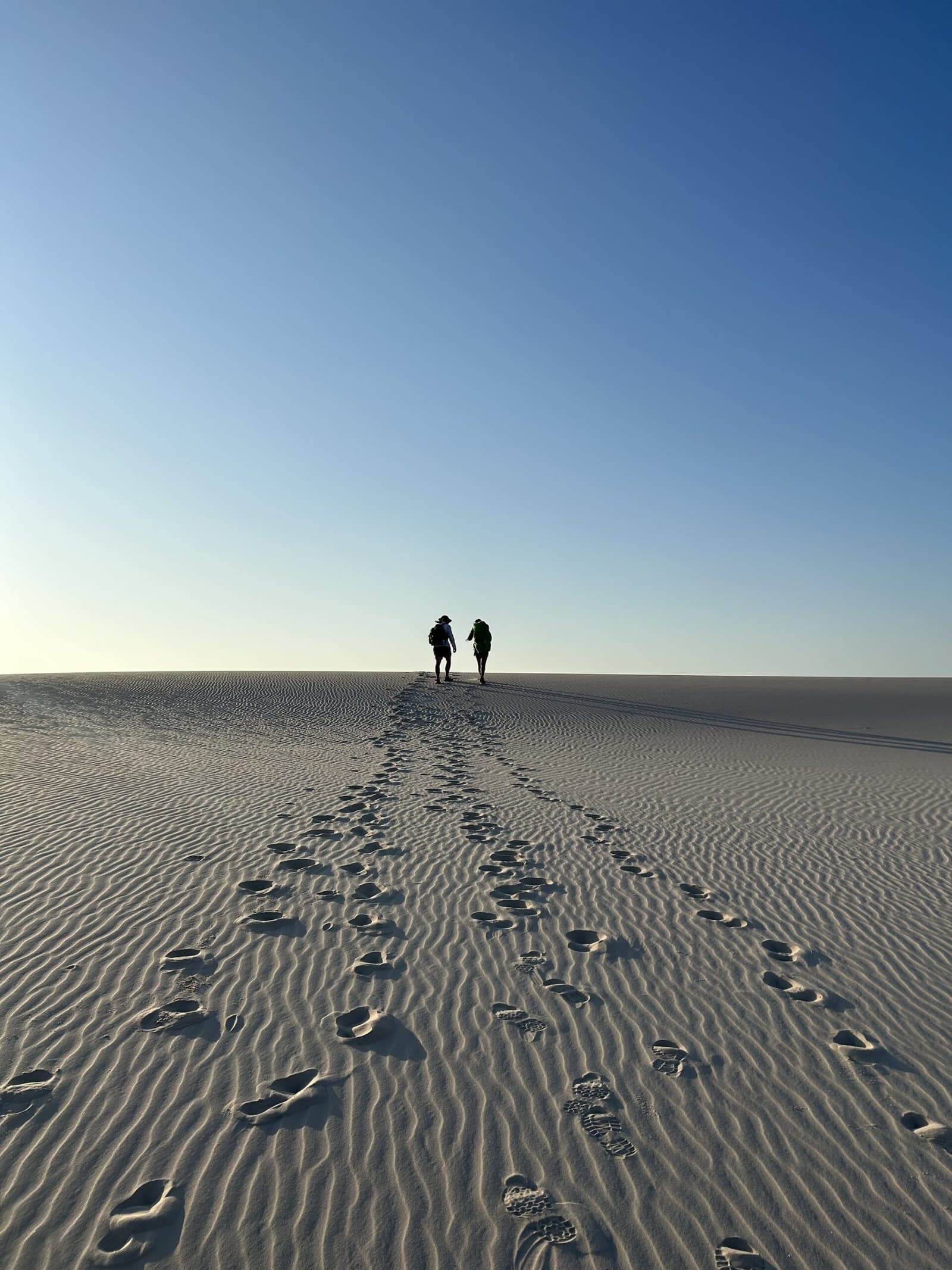 Footprints to the horizon — two trekkers crest a silent dune. Two trekkers silhouetted on the crest of a vast white sand dune during a Lençóis Maranhenses trekking expedition, a trail of footprints stretching behind them under a clear blue sky