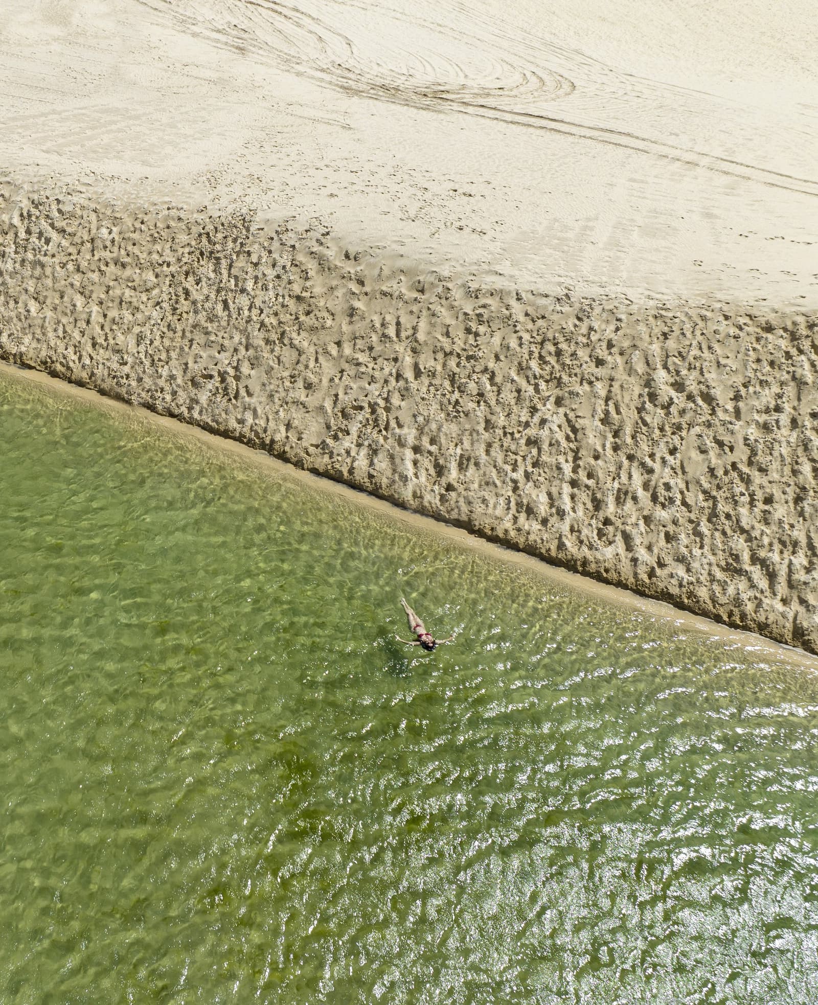 One swimmer, a vast sandbar, and water so clear you can see every ripple. Photo: Naeblys Aerial view of a lone swimmer floating in clear green water beside a sculpted sandbar during a Lençóis Maranhenses trekking experience