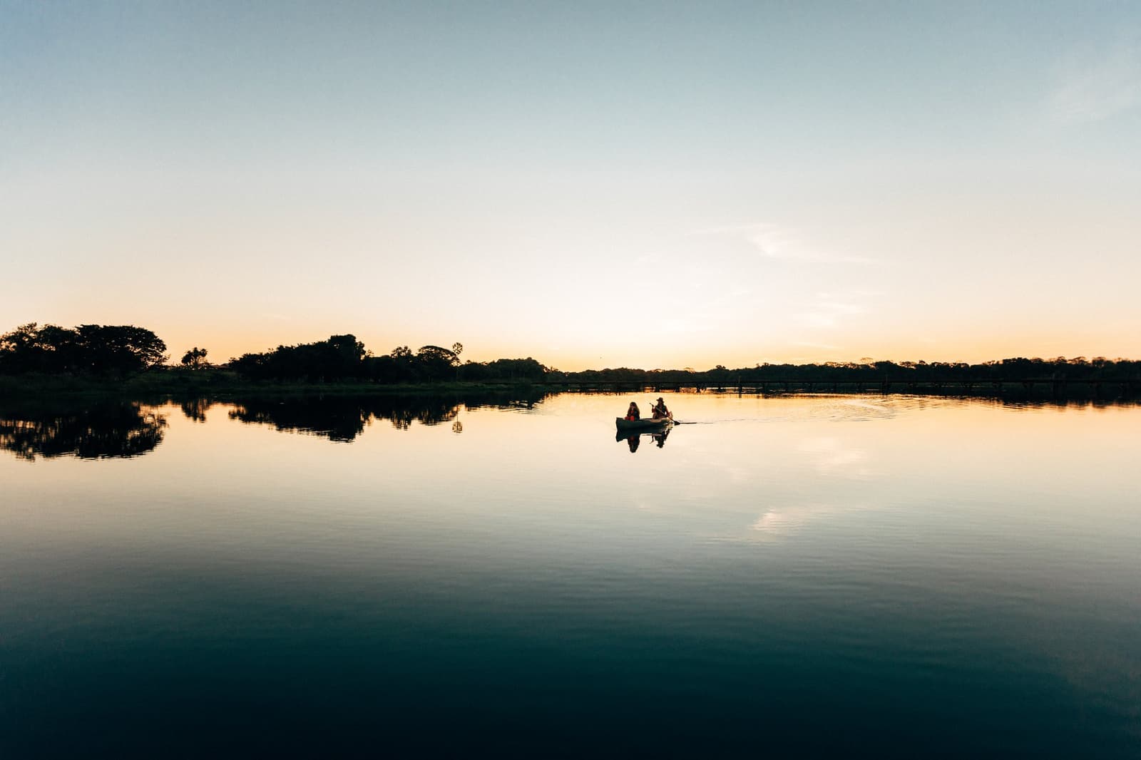 At dusk the river turns to glass, a single canoe drifting across the silence. Photo: Felipe Castellari Lone canoe with two paddlers silhouetted on a mirror-calm river at dusk on an Amazon rainforest vacation in Brazil