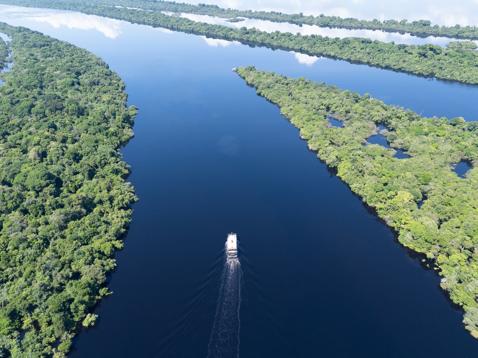 Endless green on either side, and one small boat pushing forward. Aerial view of a riverboat navigating a dark-water channel flanked by dense Amazon rainforest canopy on a luxury Amazon cruise in Brazil