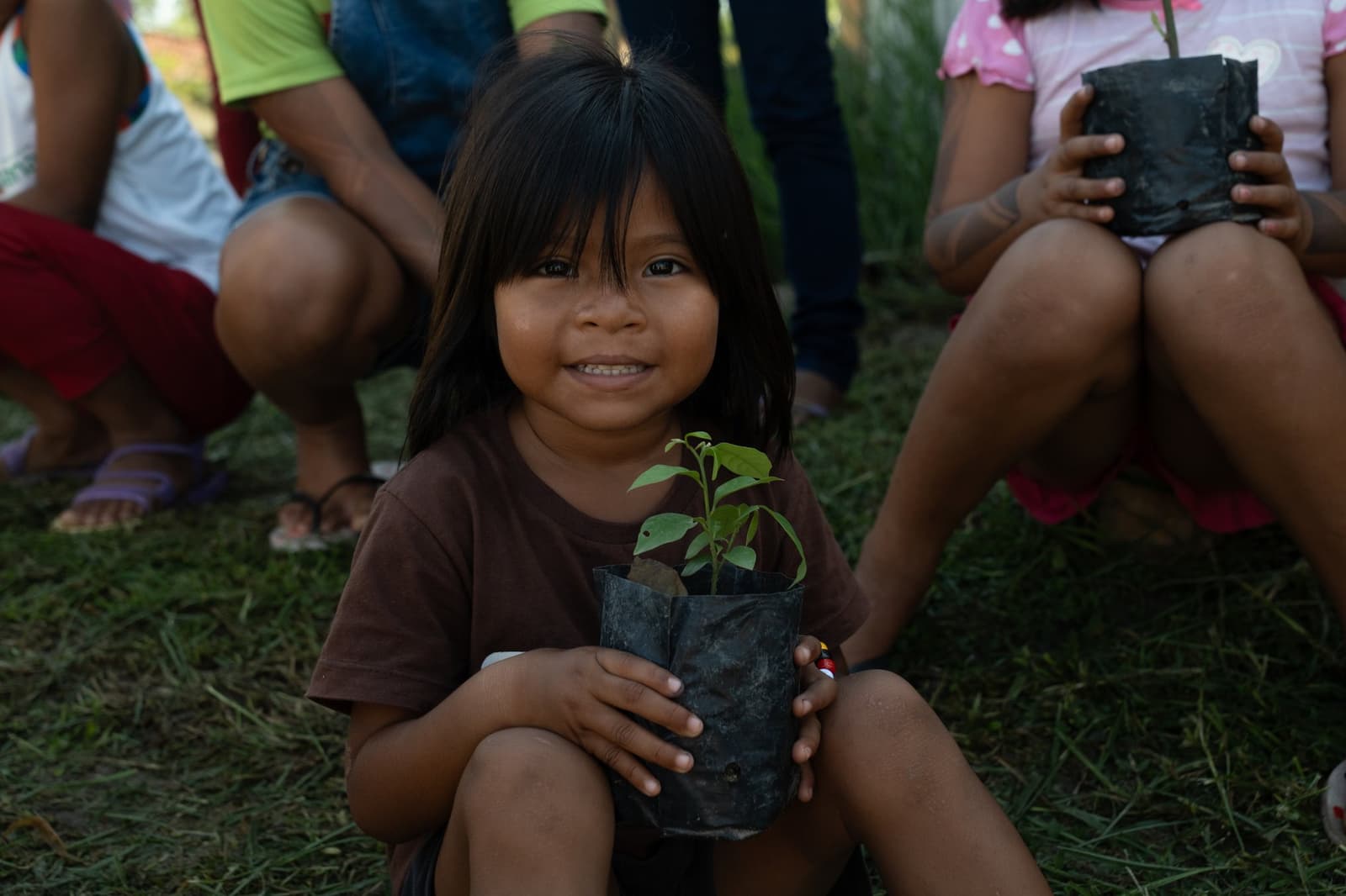 Small hands, big futures — a seedling passed between generations. A smiling young girl holds a green seedling in a black nursery pot during a community visit on a luxury Amazon cruise in Brazil, with other children seated on the grass behind her
