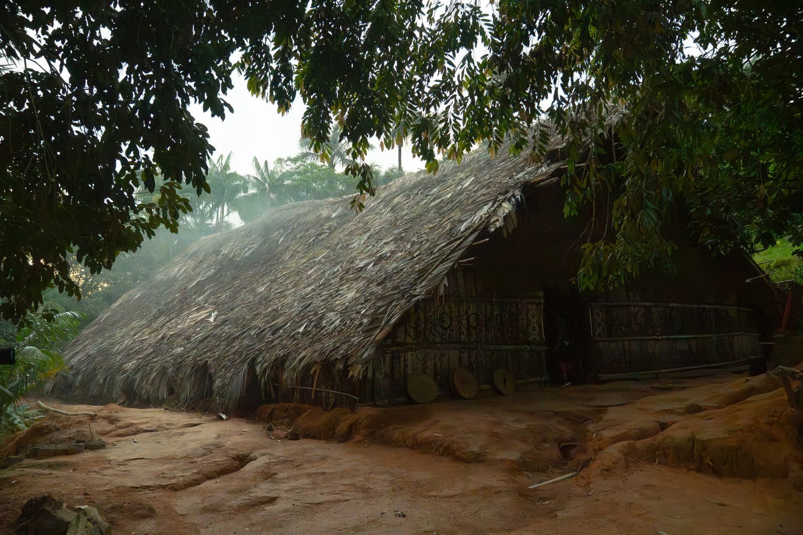 Smoke drifting past ancient rafters — a living tradition in the forest. A large palm-thatched indigenous communal longhouse with geometric painted decorations on its wooden walls, surrounded by tropical trees and red-earth ground on a Luxury Amazon Cruise in Brazil