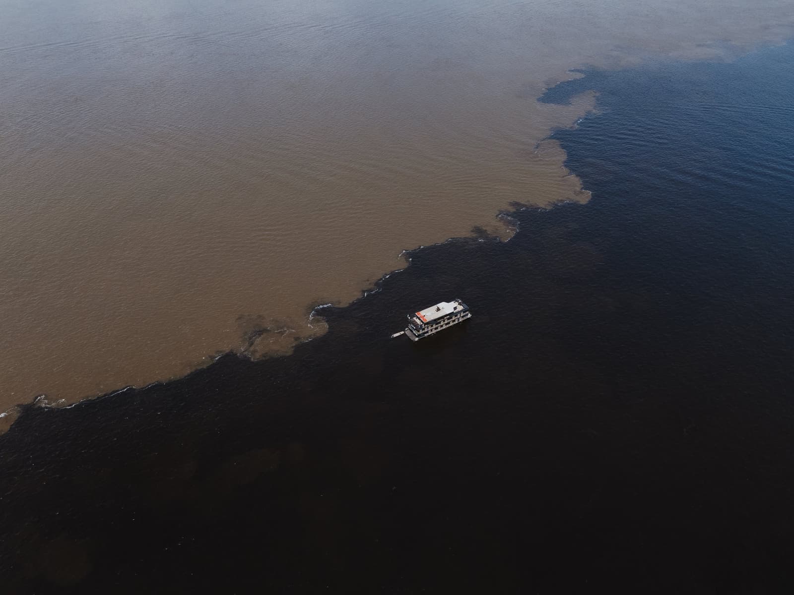 Two rivers, two colors — one line drawn by nature itself. Aerial view of a riverboat crossing the Meeting of Waters on a luxury Amazon cruise in Brazil, where dark black-water and sandy brown-water rivers meet in a sharp, winding boundary across the frame