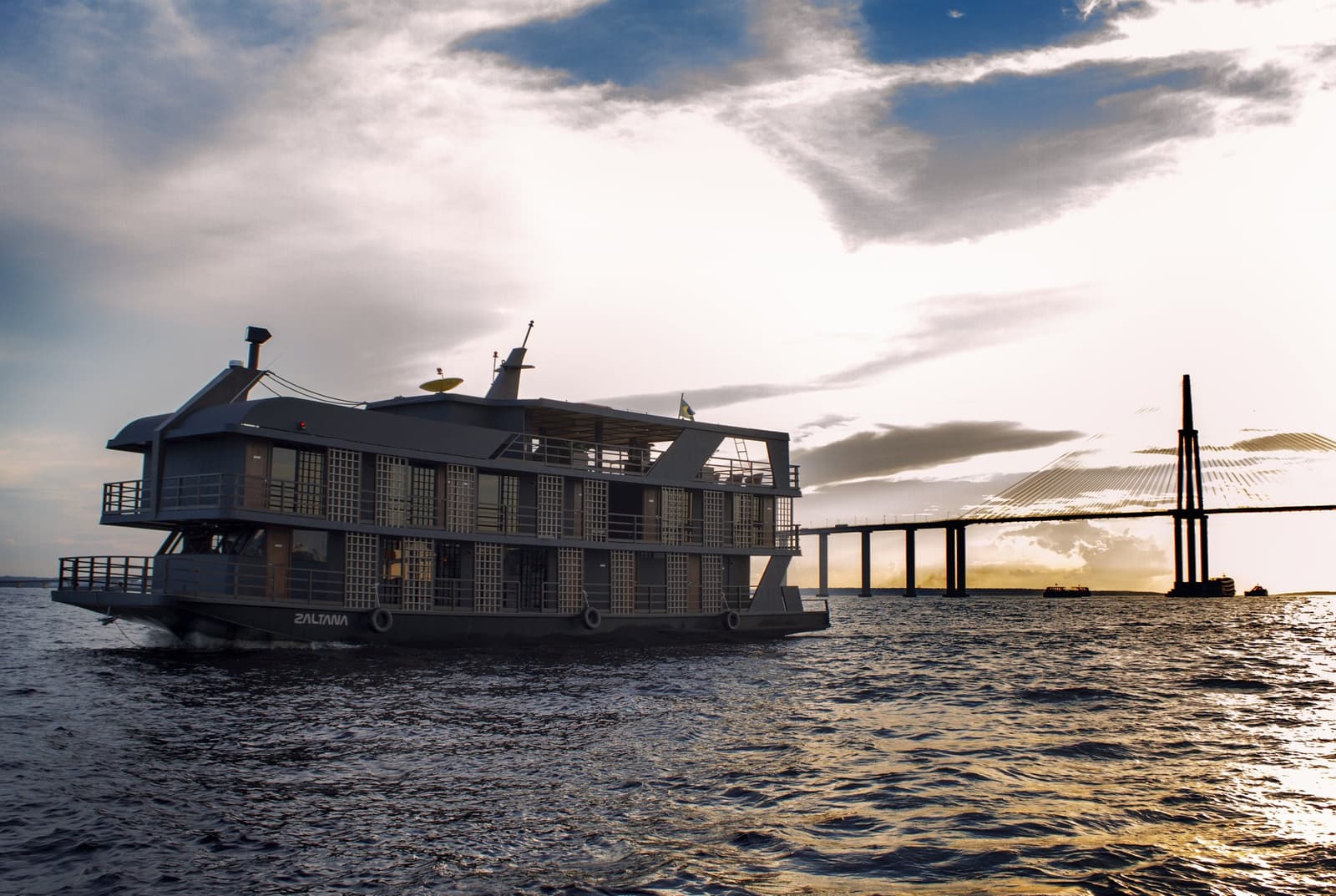 Manaus receding behind her — the Zaltana and open water ahead. The Zaltana riverboat setting out on a luxury Amazon cruise in Brazil, with a cable-stayed bridge silhouetted against a dramatic sunset sky on the broad Rio Negro