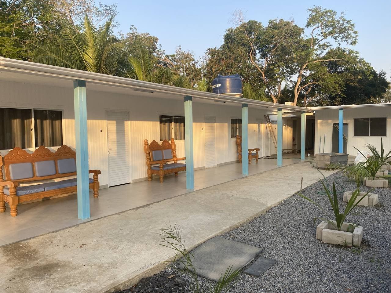 Carved benches waiting on the verandah as the afternoon light fades. A row of numbered guest rooms with carved wooden benches on a covered verandah, surrounded by tropical trees â simple, comfortable accommodation on a pantanal jaguar budget safari