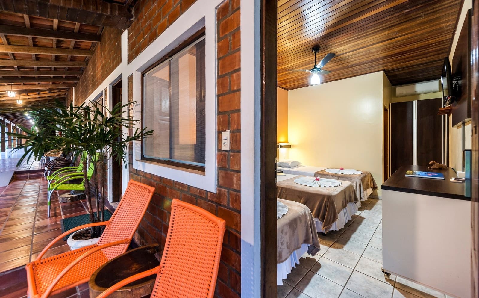 Rattan chairs on the veranda, flower-trimmed beds just beyond the door â comfort kept simple. Covered brick veranda with orange rattan chairs leading to a twin bedroom with ceiling fan and flower-decorated beds, typical of a pantanal jaguar budget safari lodge