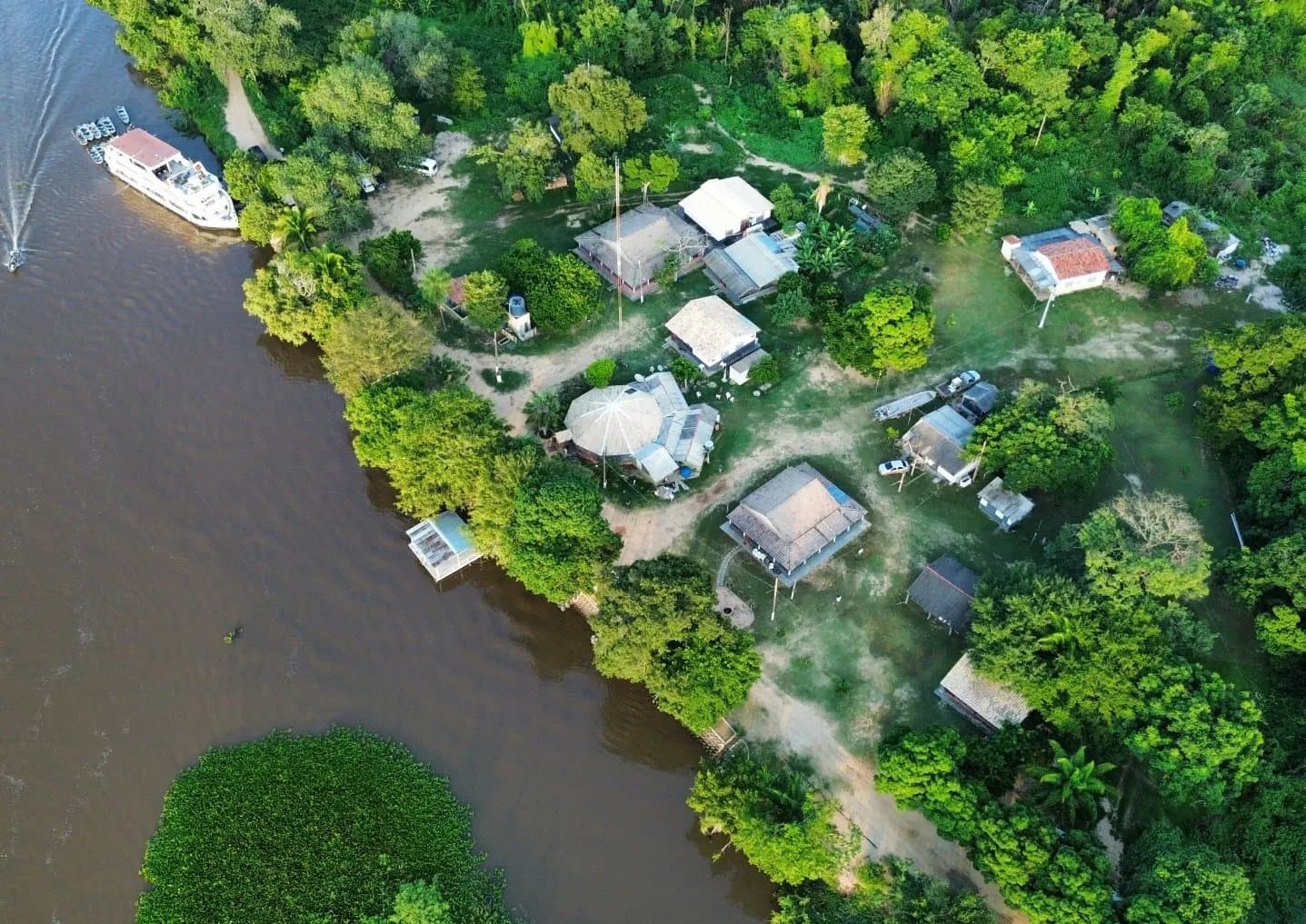 Jungle canopy on three sides, a brown river on the fourth â your base for a Pantanal jaguar budget safari. Aerial drone view of a small riverside lodge tucked into dense green forest along a brown river in the Pantanal, with a multi-deck boat moored at the bank and thatched-roof buildings set among tropical trees