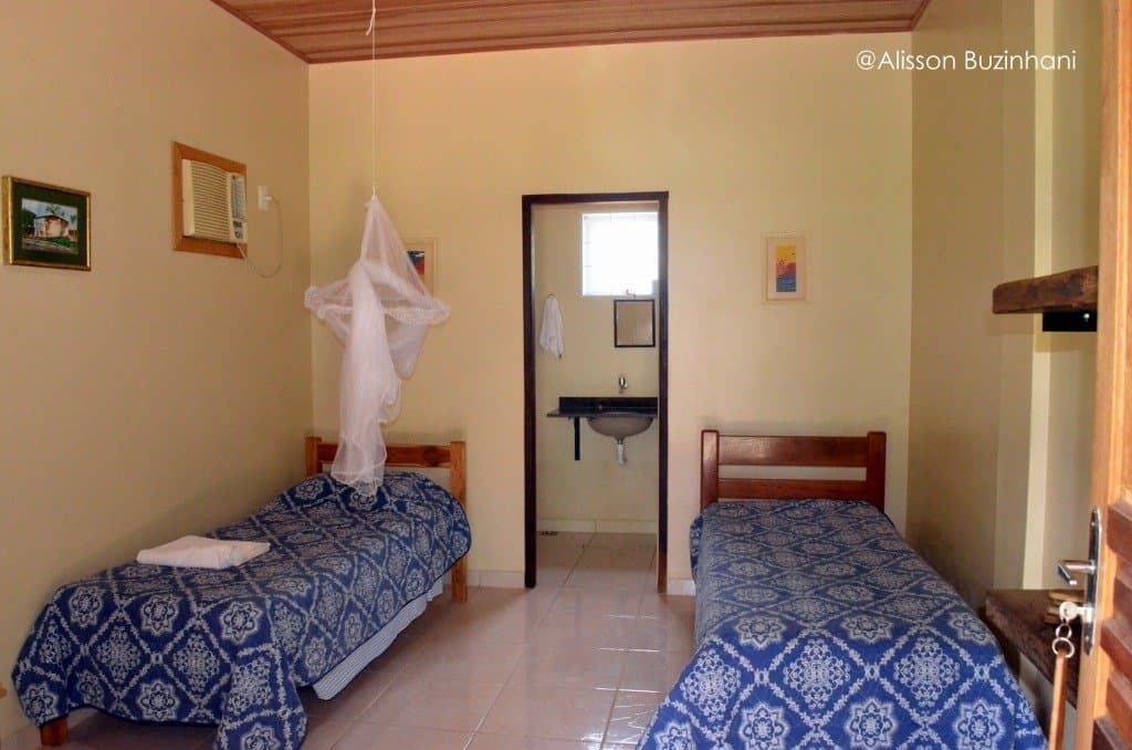 Mosquito net overhead, matching beds ready â simple comfort after a long day on the river. Twin bedroom with blue patterned bedspreads, a hanging mosquito net, and an en-suite bathroom visible through the doorway at a simple Pantanal jaguar budget safari lodge