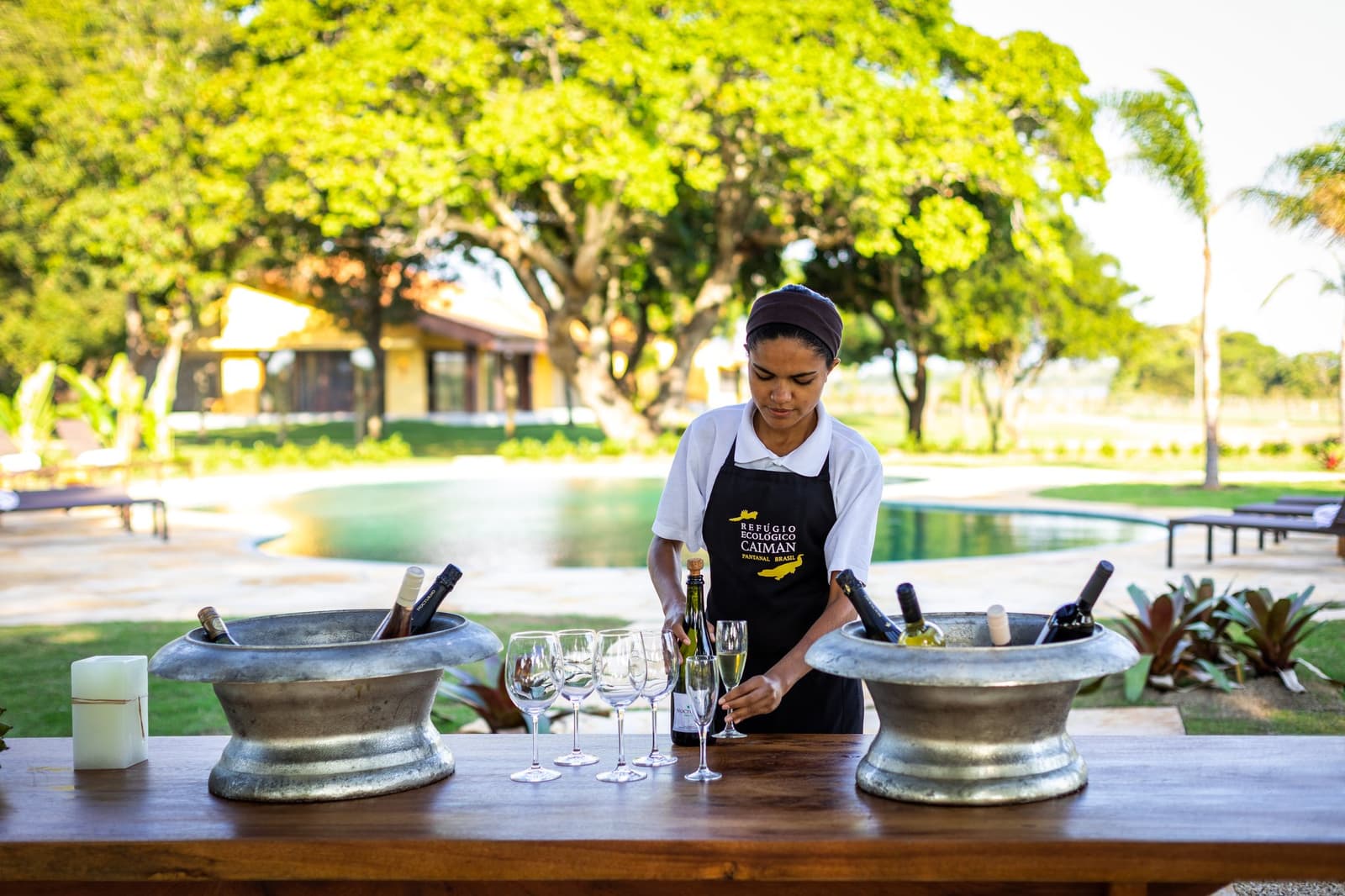 Sparkling wine, silver ice buckets, and a warm welcome waiting at the bar. A Caiman Ecological Refuge staff member in a branded apron arranging sparkling wine glasses and chilled bottles in metal ice buckets on a wooden bar, with a swimming pool and lush grounds visible behind her — part of the pantanal luxury safari experience