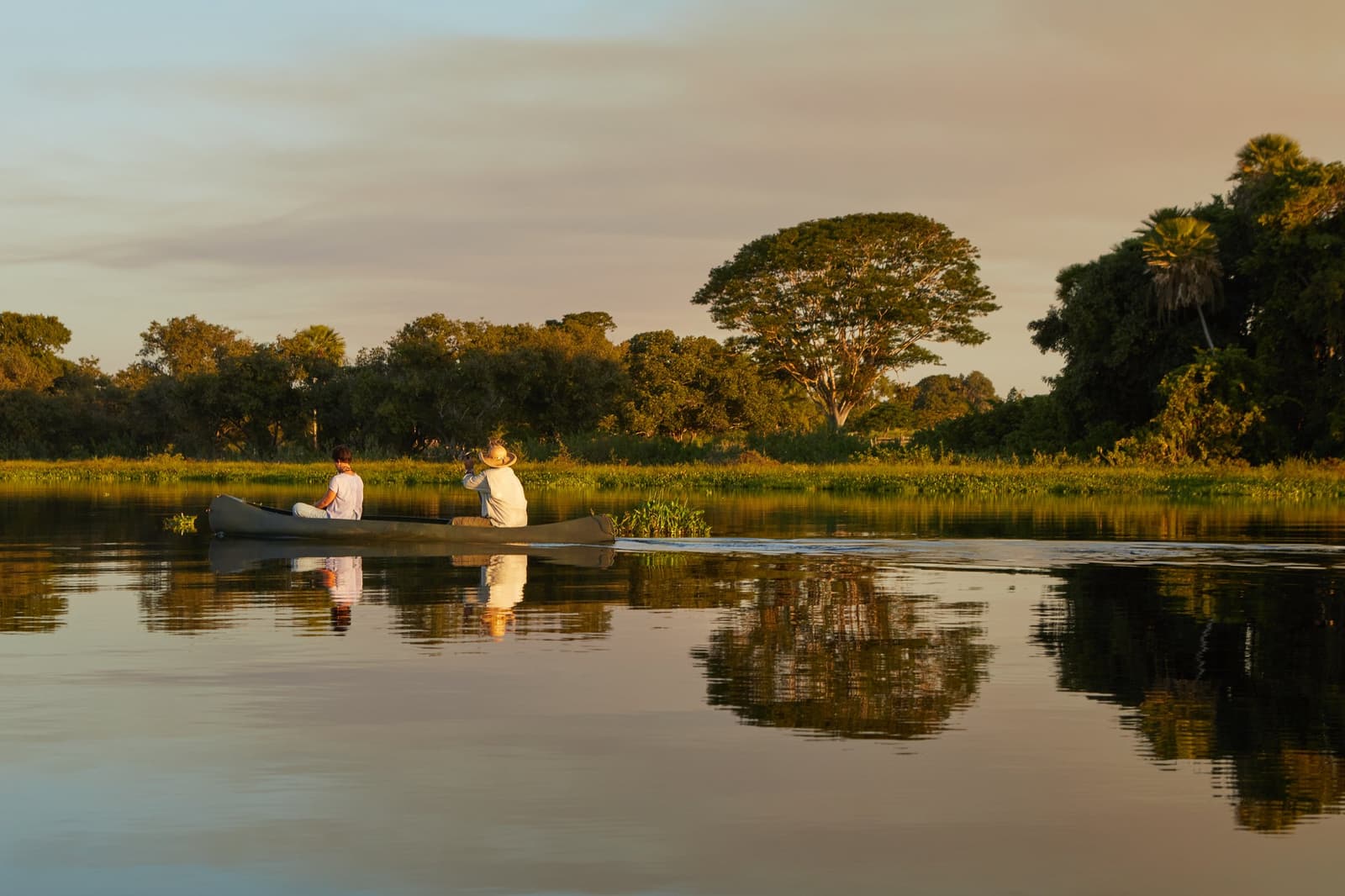 Paddling softly into the last light of the day. Two guests drifting in a canoe on glassy water at golden hour during a pantanal luxury safari, surrounded by lush wetland vegetation and large trees reflected in the still river