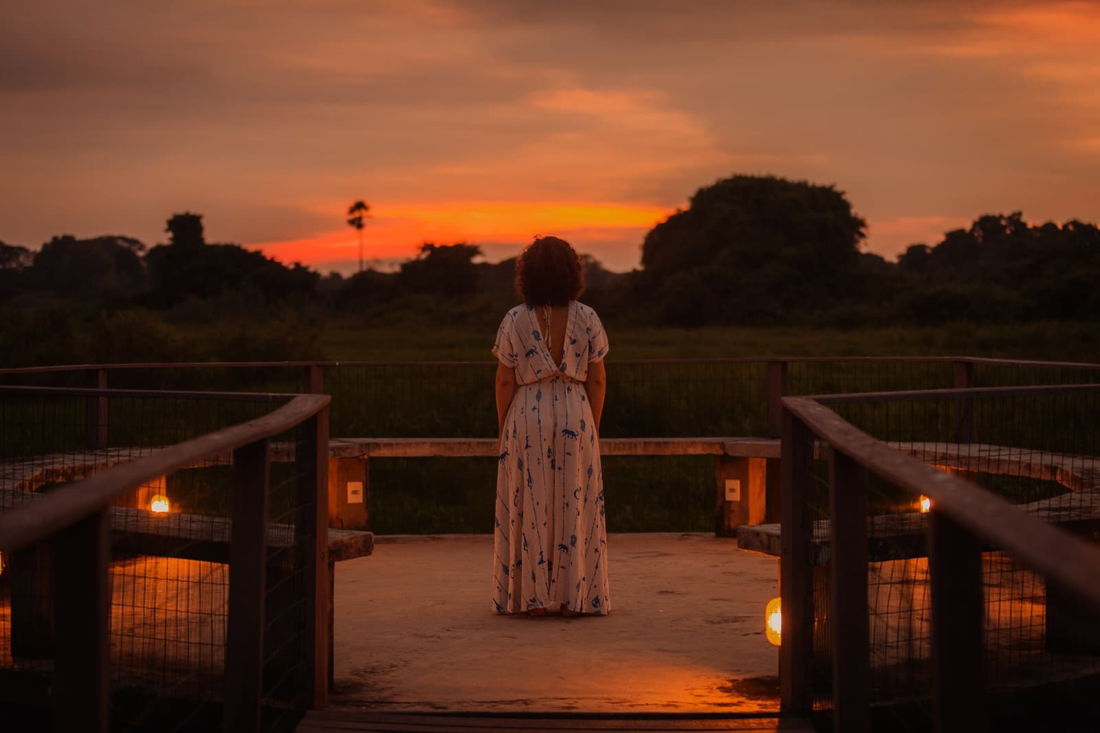 Still skies, burning horizons — evenings here belong entirely to you. A woman in a floral maxi dress stands on a candlelit wooden deck, watching a blazing orange sunset over open wetland flats on a pantanal luxury safari